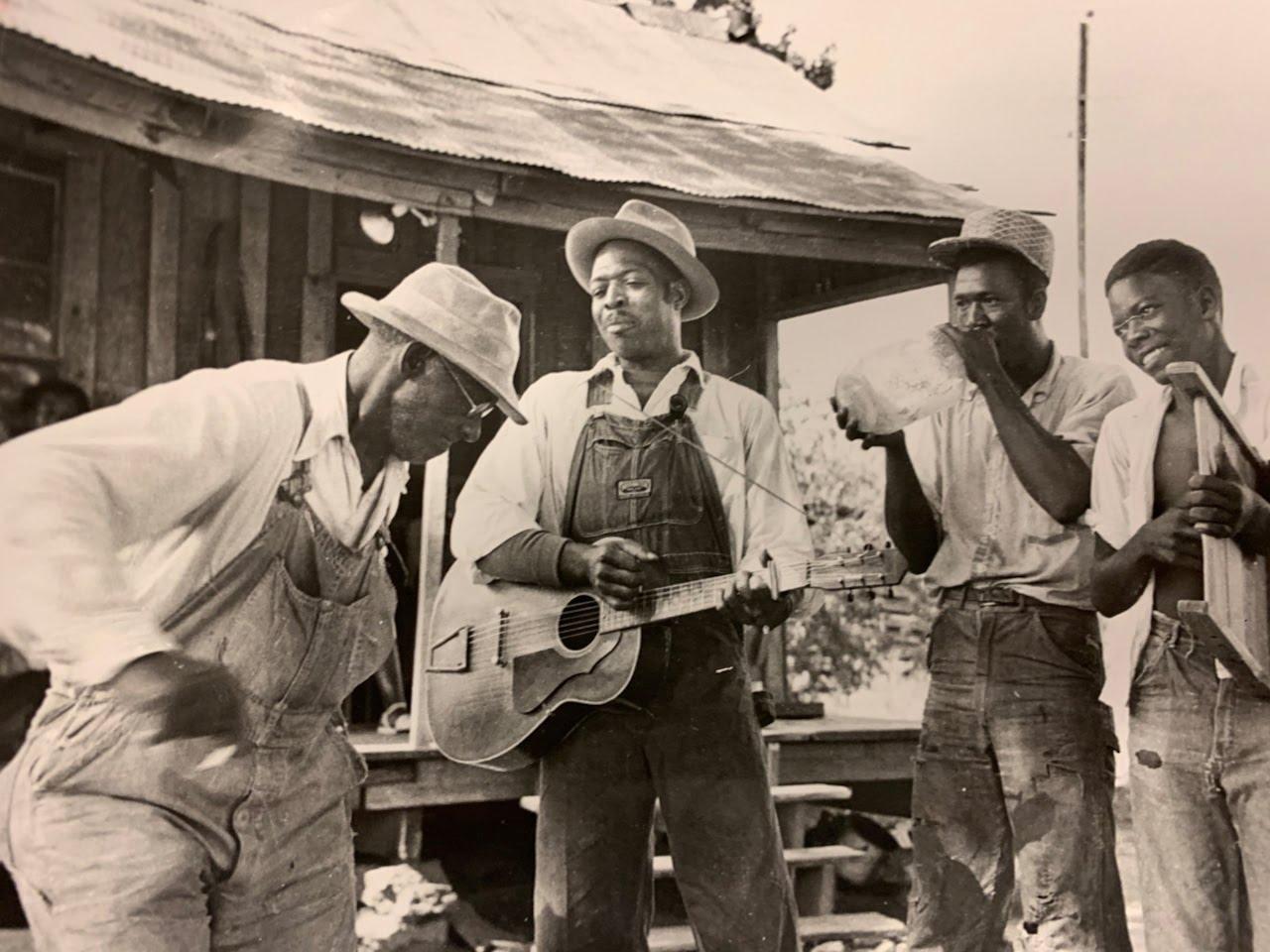 Black and white photo of  a jug band. There is a guitar player, someone playing a jug and a man dancing.