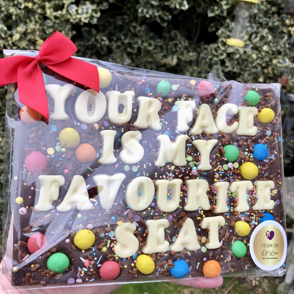 Large novelty chocolate slab with white chocolate lettering reading ‘Your face is my favourite seat’, decorated with colourful candy pieces and sprinkles, wrapped with a red ribbon.