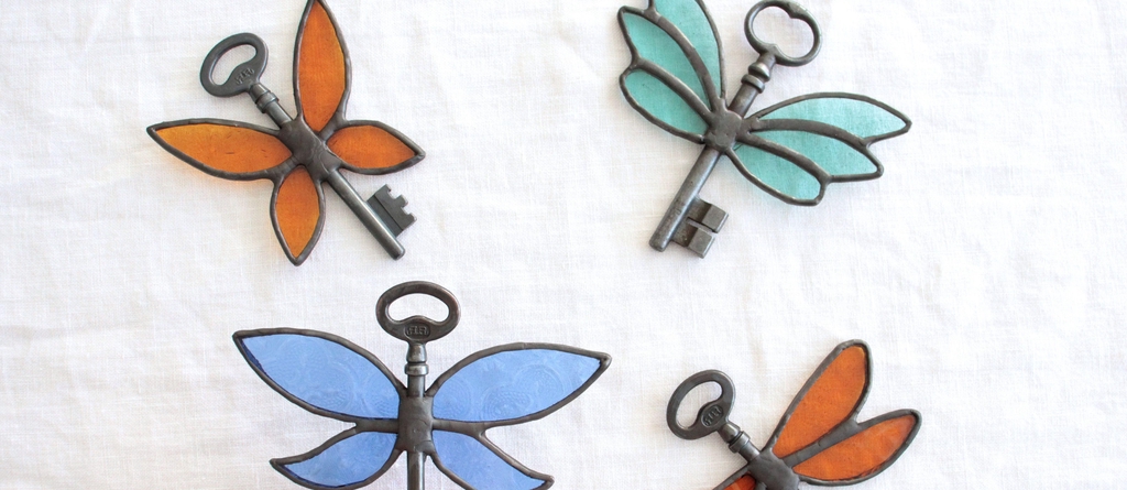 Photo de Atelier Vitraï montrant quatre clés noires ornées d'ailes en vitrail bleues, oranges et vertes.