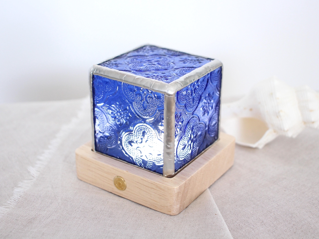 Photo de Atelier Vitraï montrant une lampe en vitrail bleu en forme de cube, sur son socle en bois clair.
