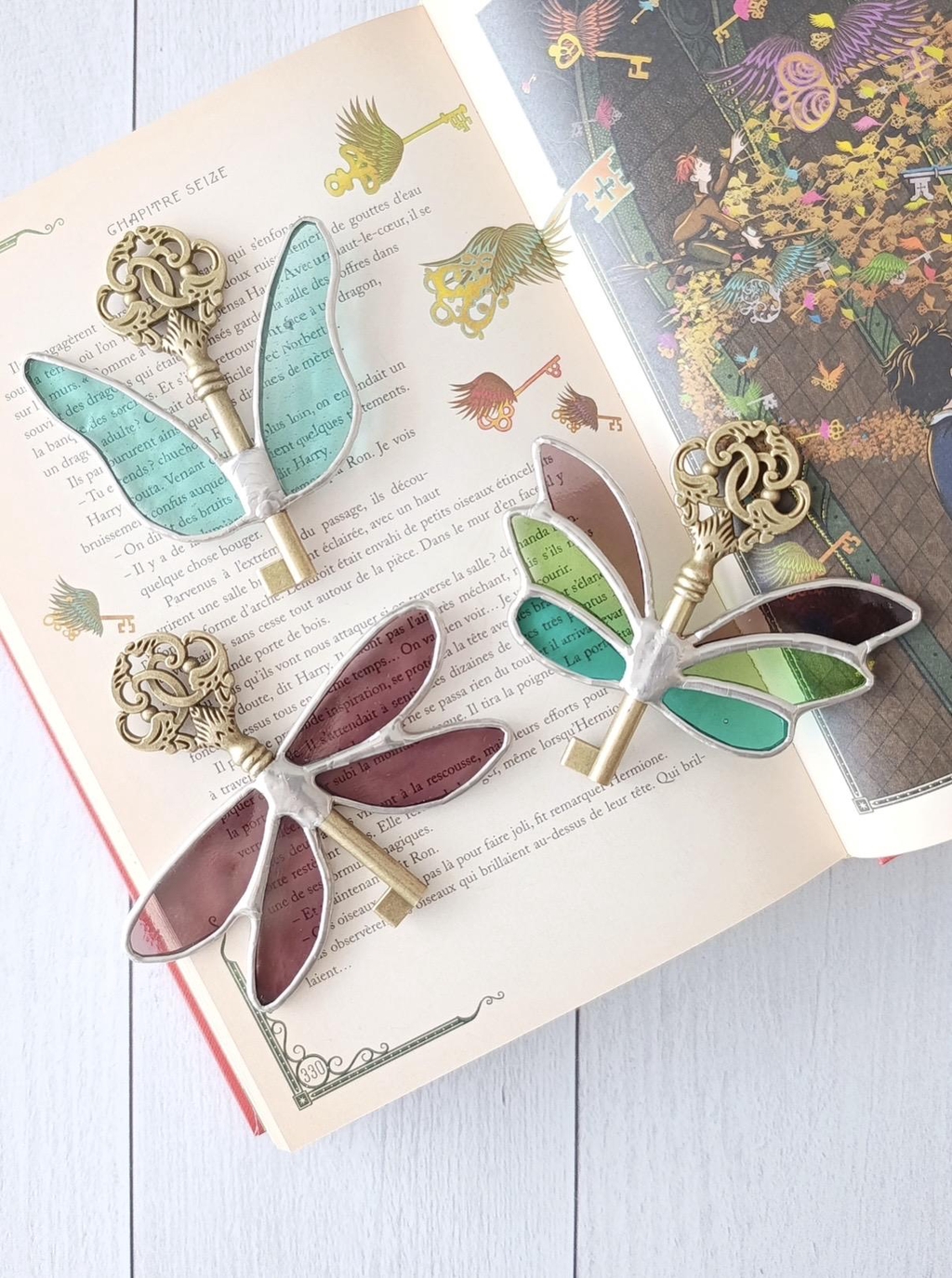 Photo de Atelier Vitraï montrant trois clés dorées ornées d'ailes en vitrail bleues, vertes et violettes.