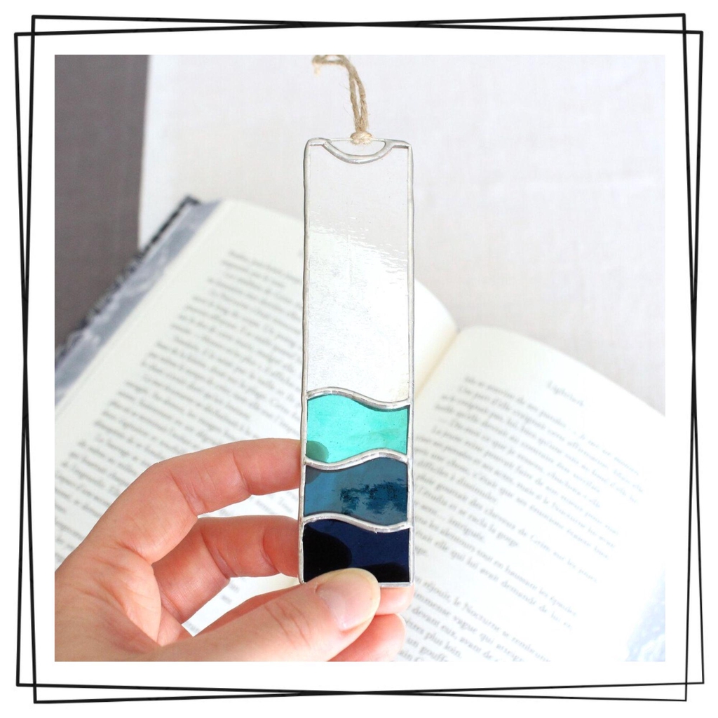 Photo de Atelier Vitraï, montrant un marque-page en vitrail arborant des vagues bleues, tenu par une main au-dessus d'un livre.