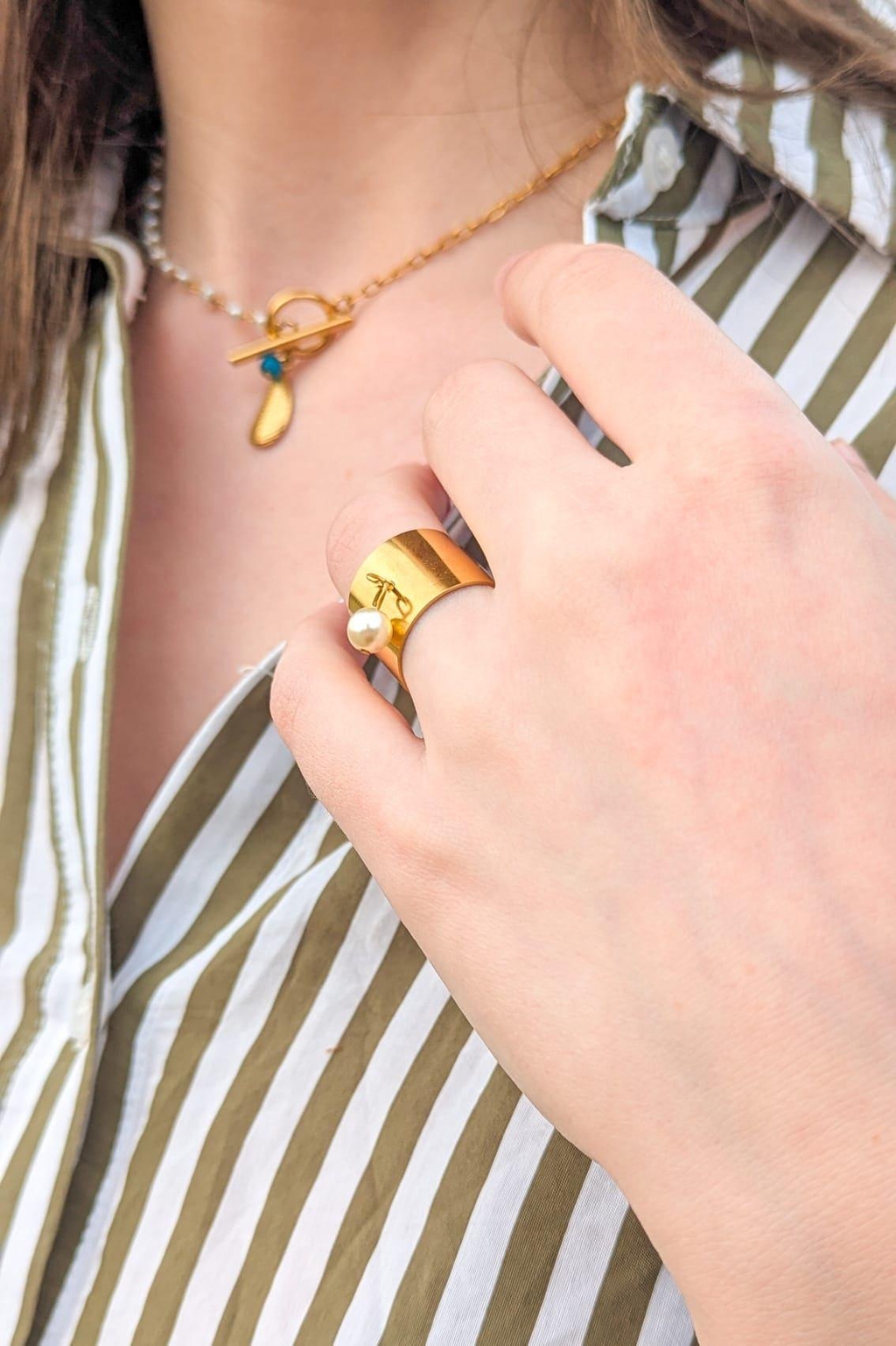 Bague ajustable dorée avec perle portée au doigt, collier chaîne dorée en arrière-plan – bijoux fantaisie en acier inoxydable Les Althëis