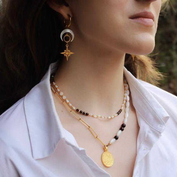 Femme portant des bijoux dorés Les Althëis : boucles d'oreilles pendantes croissant de lune et étoile polaire avec perle blanche en nacre, et deux colliers superposés - un rang de perles blanches et rouges, et une chaîne dorée avec médaillon gravé ovale du disque de Phaistos.