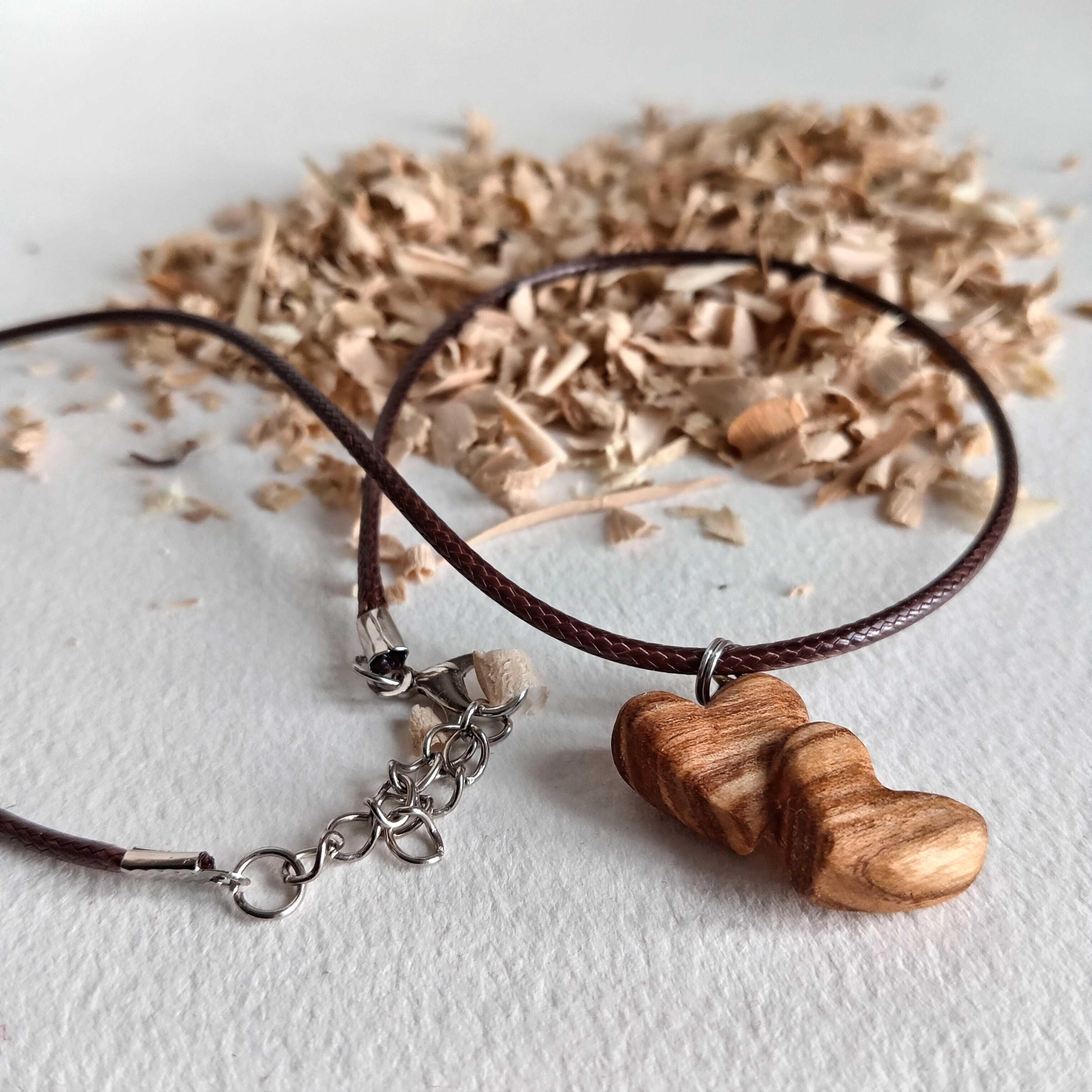 Pendentif en bois de frêne olivier représentant deux cœurs entremêlés accrochés à un collier en coton ciré posés sur des copeaux de bois.