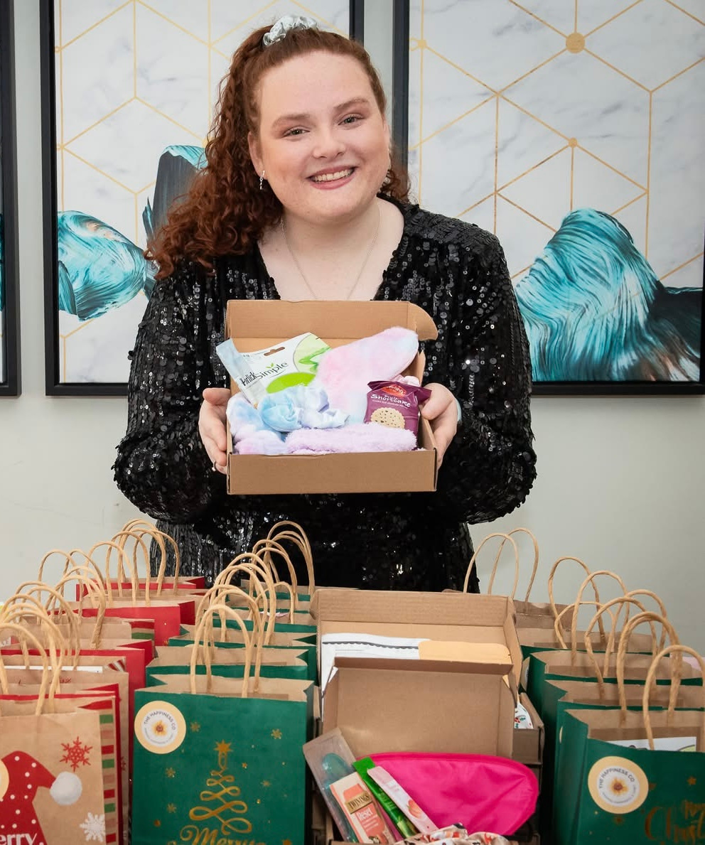 A smiling woman with curly hair, wearing a black sequin dress, holds open a small self-care gift box behind a table filled with holiday gift bags.