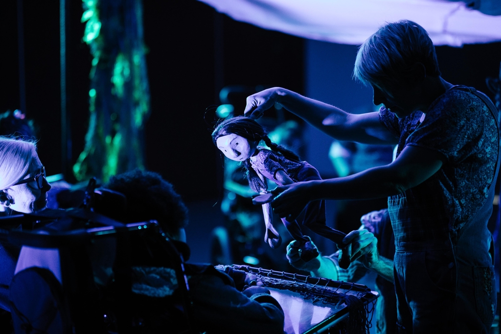 A photo taken during a Bamboozle performance. There is a puppet of a woman with plaited hair looking at a young person in a wheelchair.
