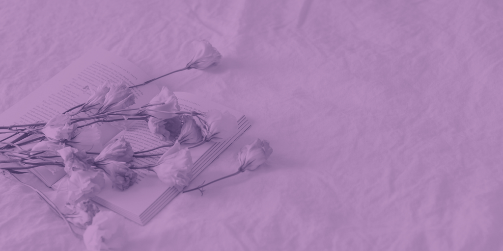 Un fond dans les tons mauves avec un livre ouvert et des fleurs posés sur un drap. 