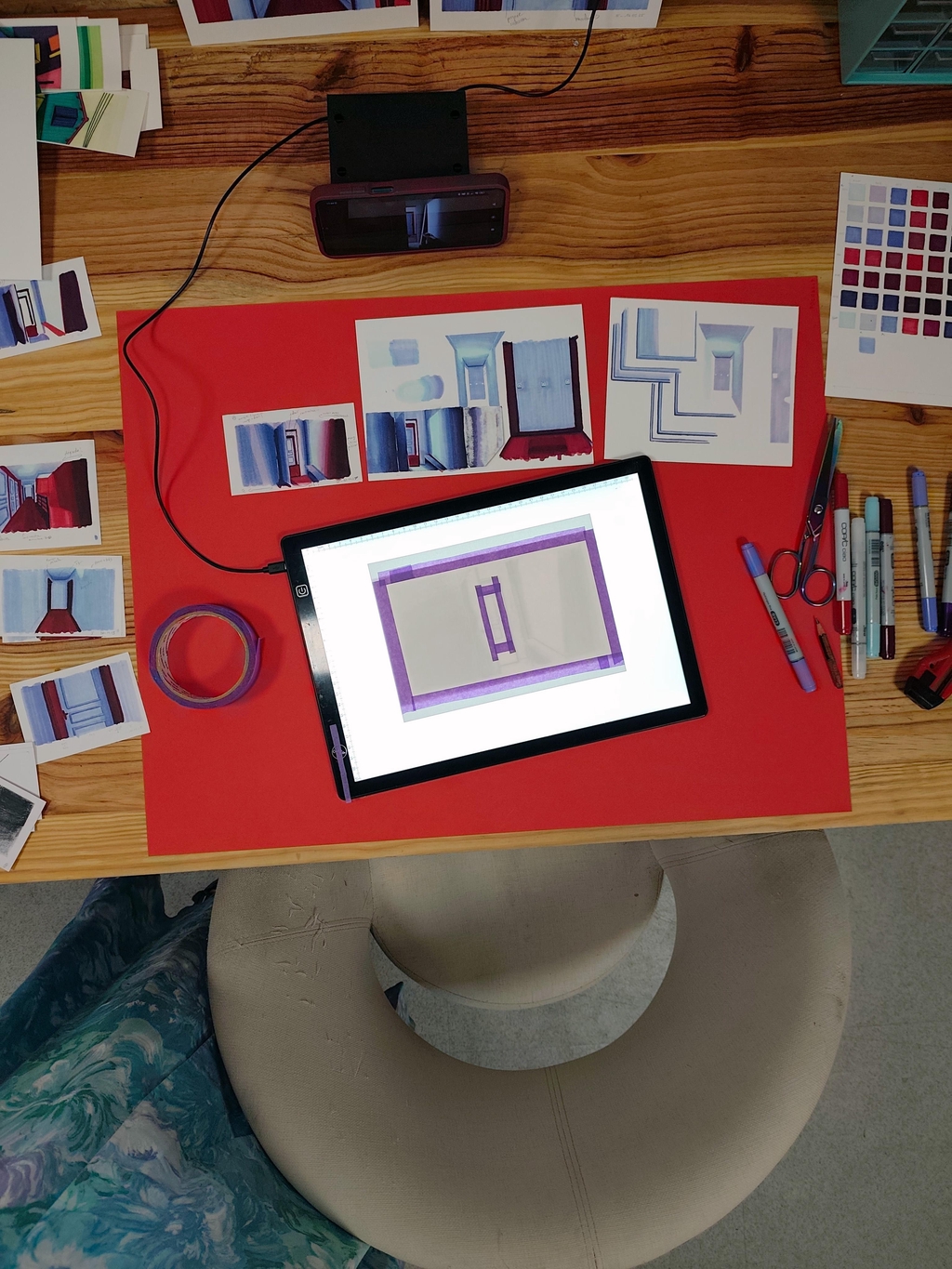 Photo de la table de travail vue de haut. Au centre une tablette lumineuse sur une grande feuille rouge. Les essais d'encrages et feutres tout autour.