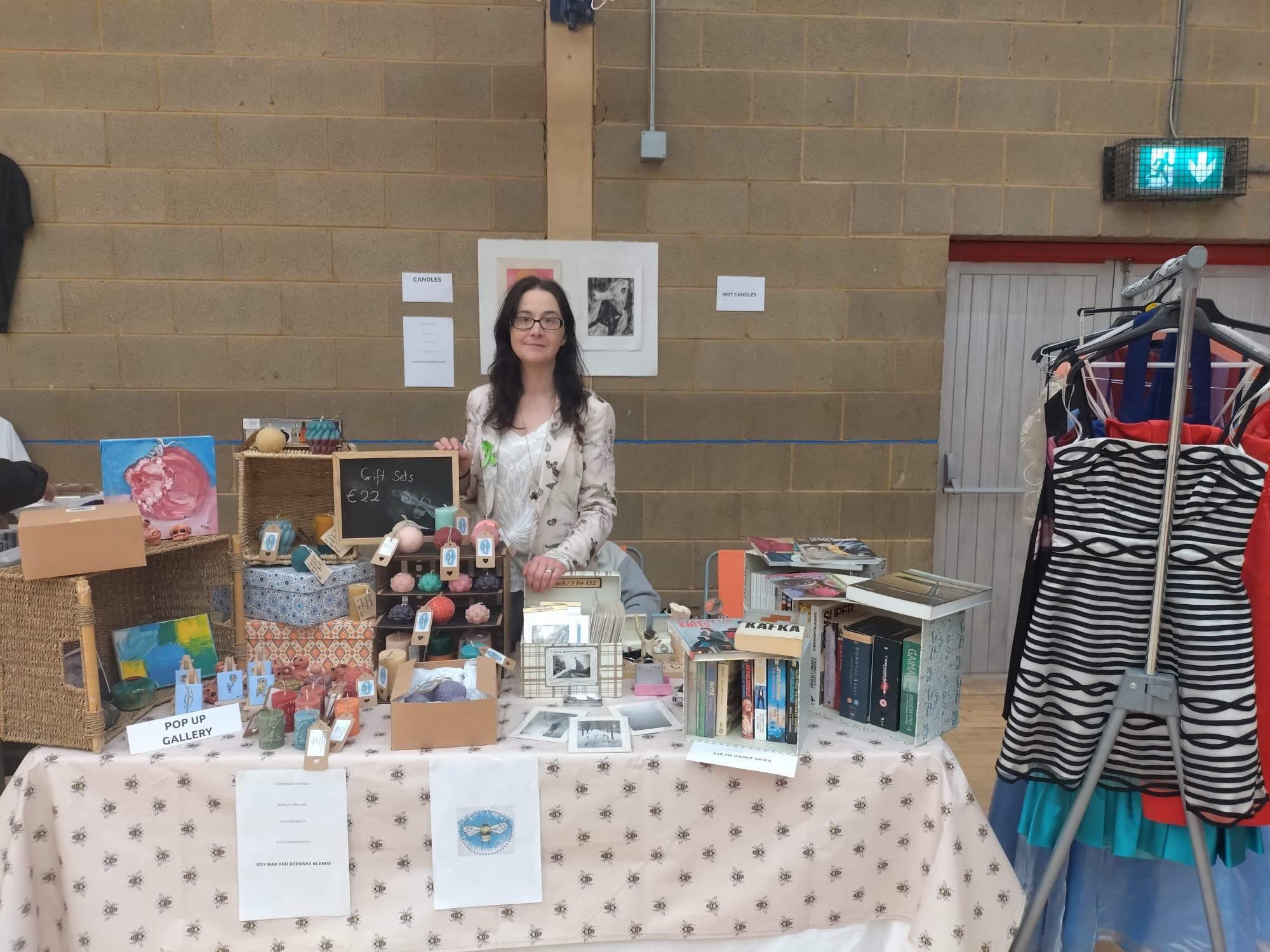 Person standing behind a stall. There are wide range of items on the stall. Candles, Art, hobbies, books. The corner of the right hand side of the photo has part of a clothes rail the rest is not visible.