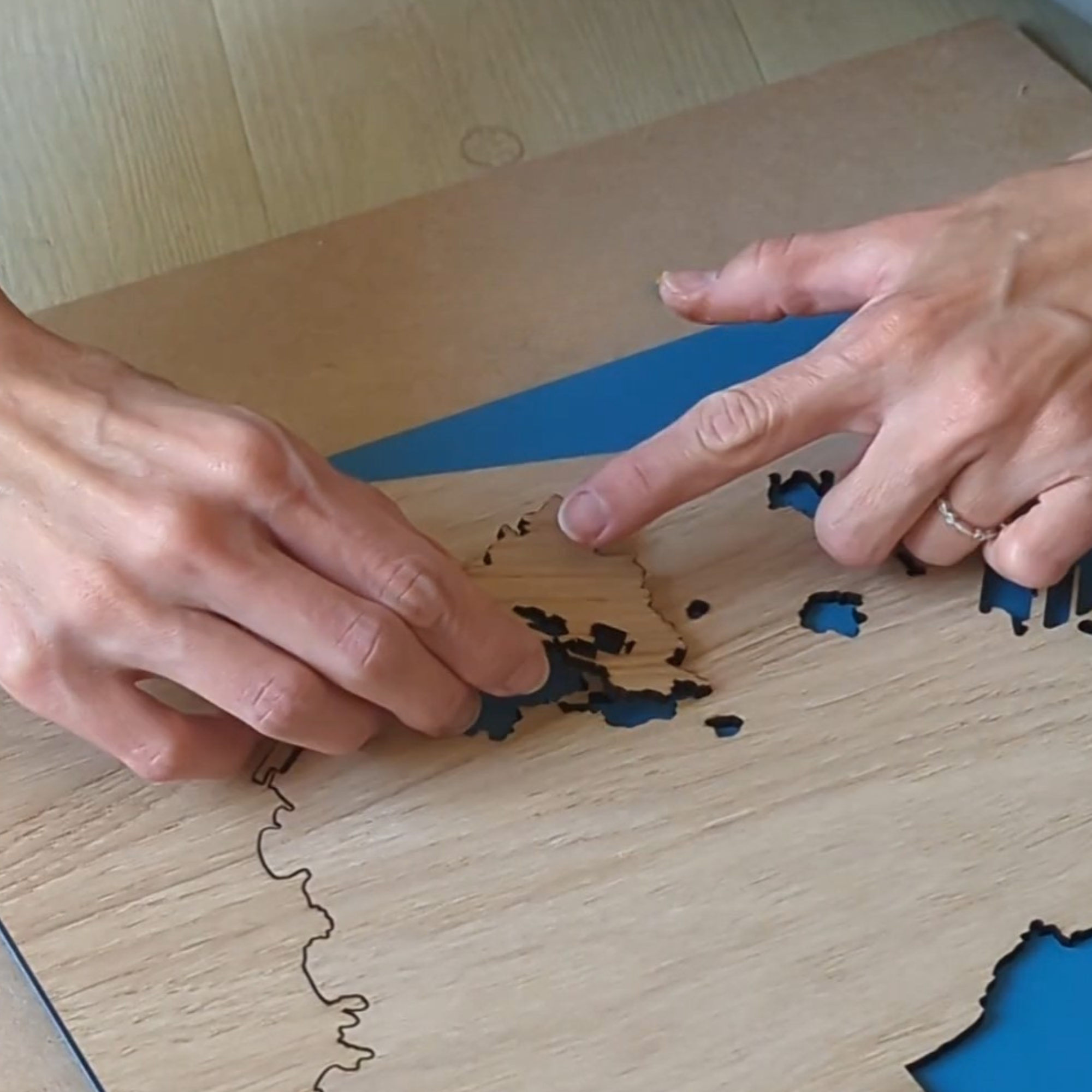 Fabrication d’un tableau cartographique Litto-art : mains assemblant une pièce en bois de chêne représentant une île sur un fond bleu.