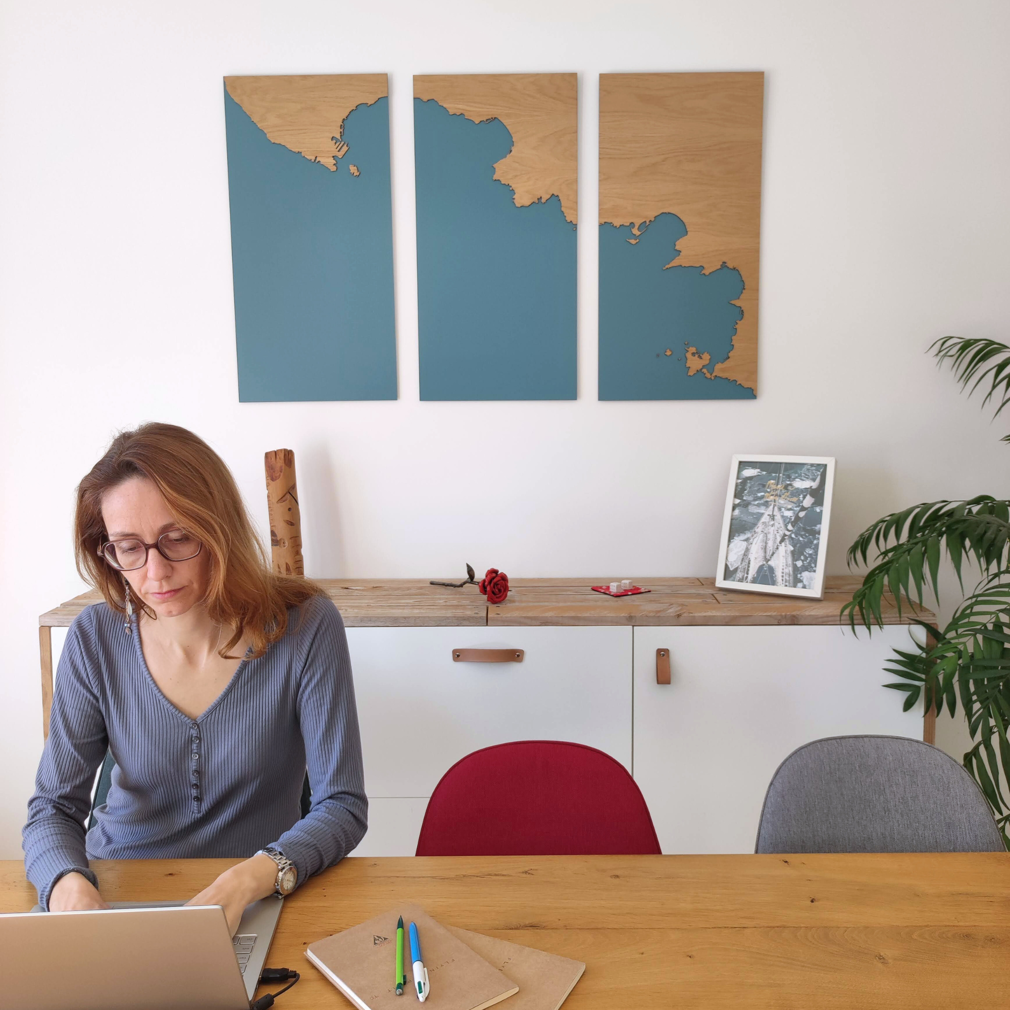 Marion, créatrice de Litto-art, travaillant sur ordinateur devant un grand tableau cartographique en bois représentant le littoral, dans un intérieur décoré de façon naturelle et artisanale.