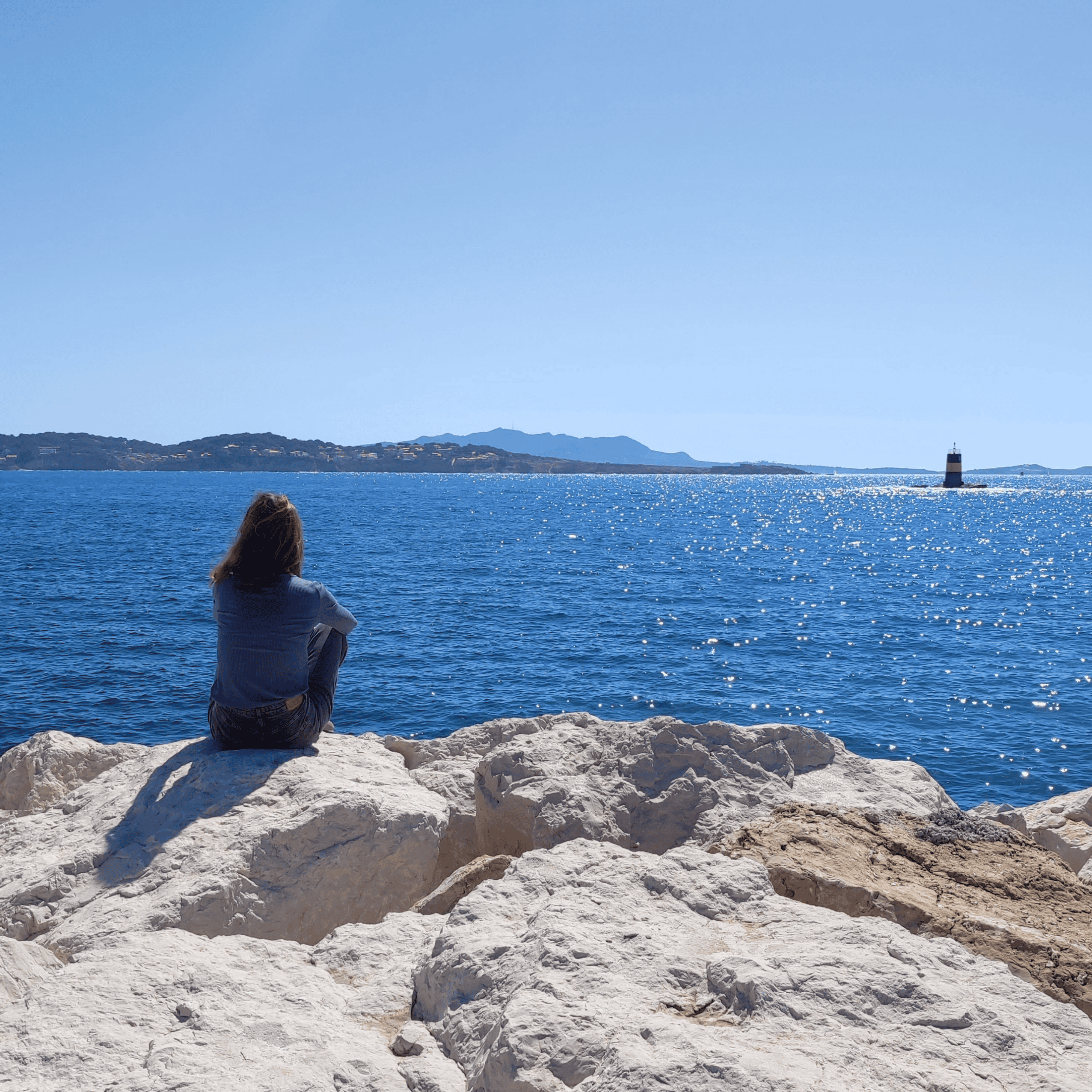 Créatrice de Litto-art assise sur une digue en enrochements face à la mer Méditerranée et regardant le littoral varois.