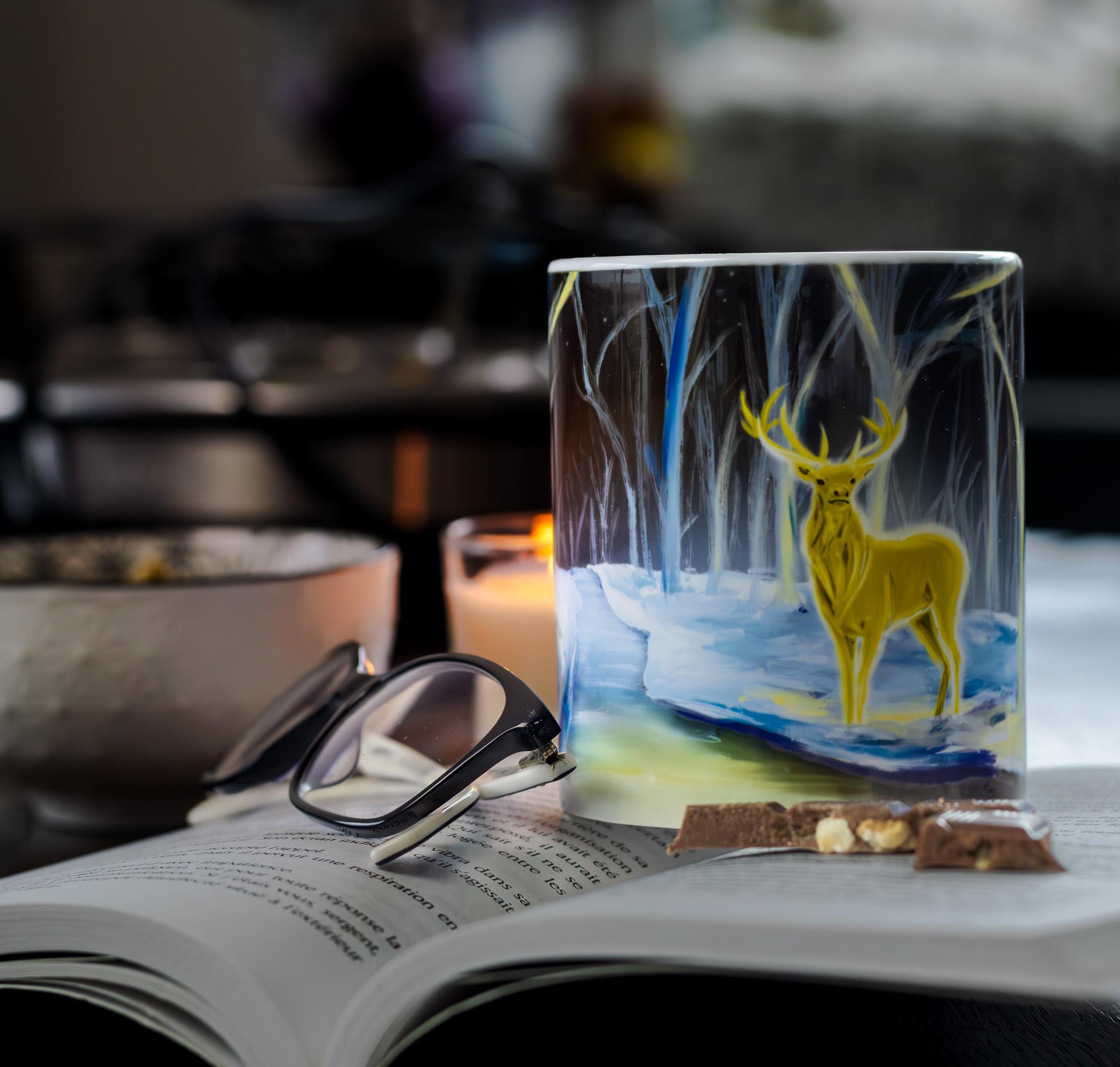 Tasse d'un cerf d'oré dans la nuit bleuté enneigé. Ambiance cosy. Sur un livre avec du chocolat et une lueur d'une bougie au loins
