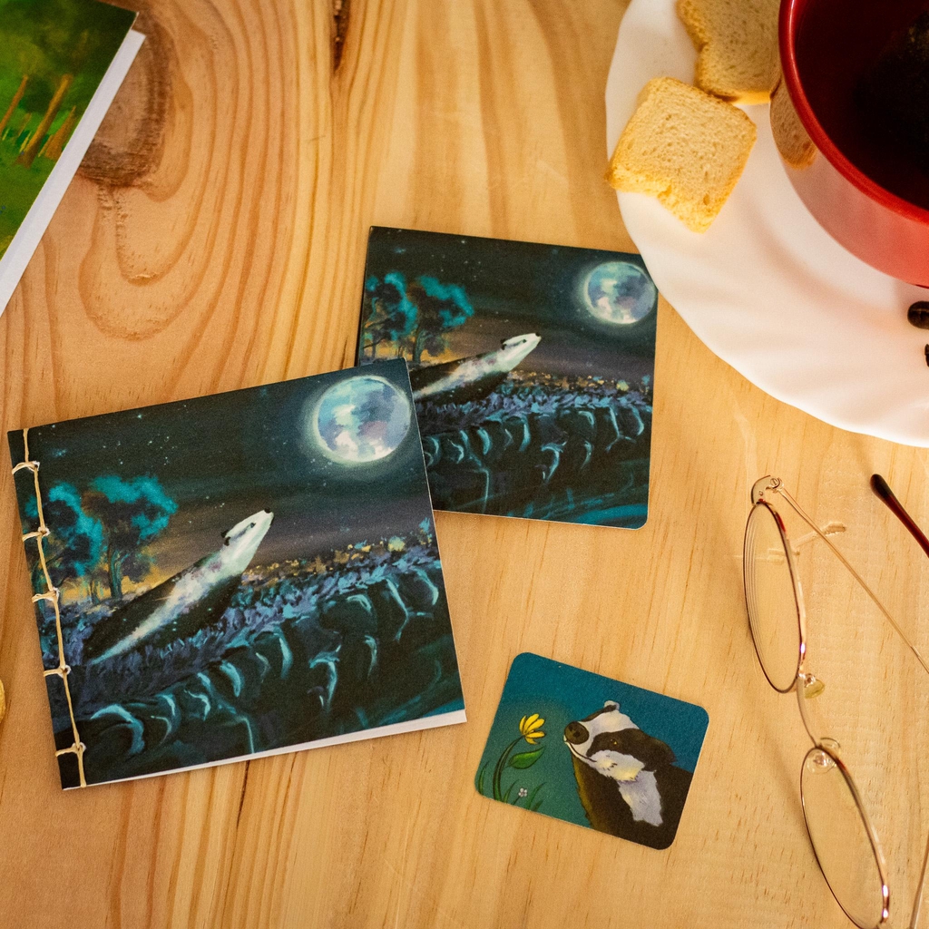 Petit livre 10x10 relier à la main sur une table en bois à coter d'une tasse de thé, moment cosy. Livre illustré d'un blaireau au pelage étoilé regardant la lune