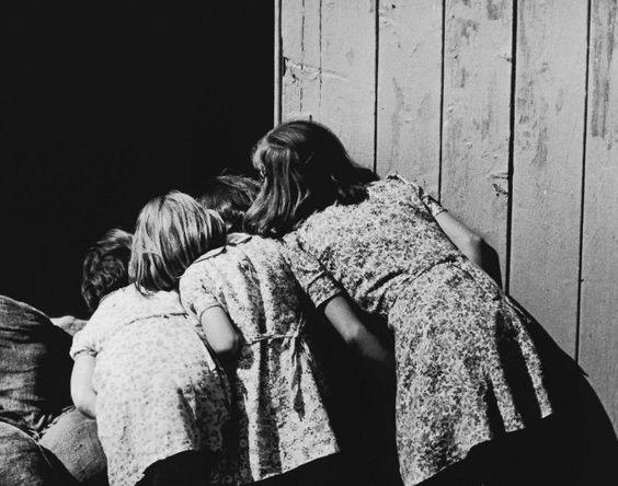 Photo en noir et blanc vintage, des années 60, 70. La photographie représente 4 petites filles de dos, en robe fleuri, qui se cache en regardent derrière une porte en bois. 