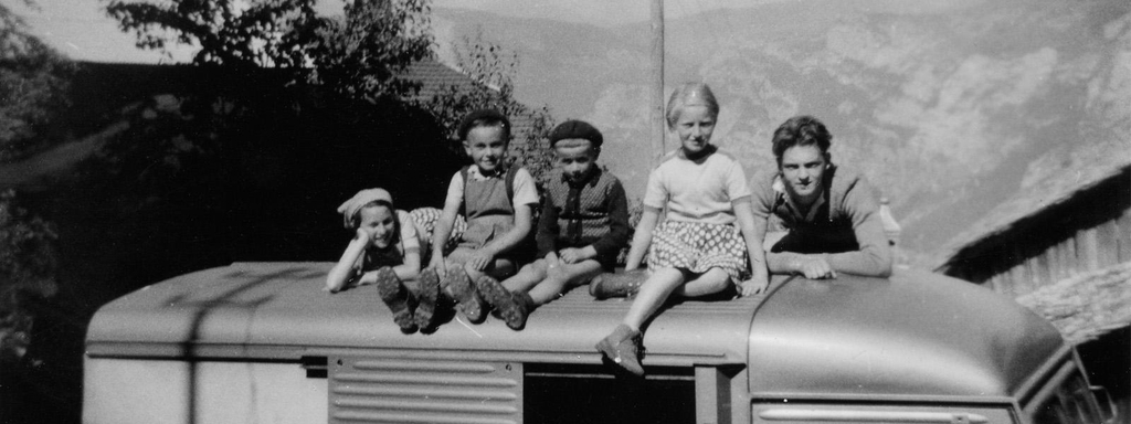 Photo en noir et blanc des années 40 - 50. La photographie représente 5 enfants sur le toit d'une camionnette avec en arrière-plan les montagnes de Haute Savoie. Il y a 2 filles et 3 garçons.