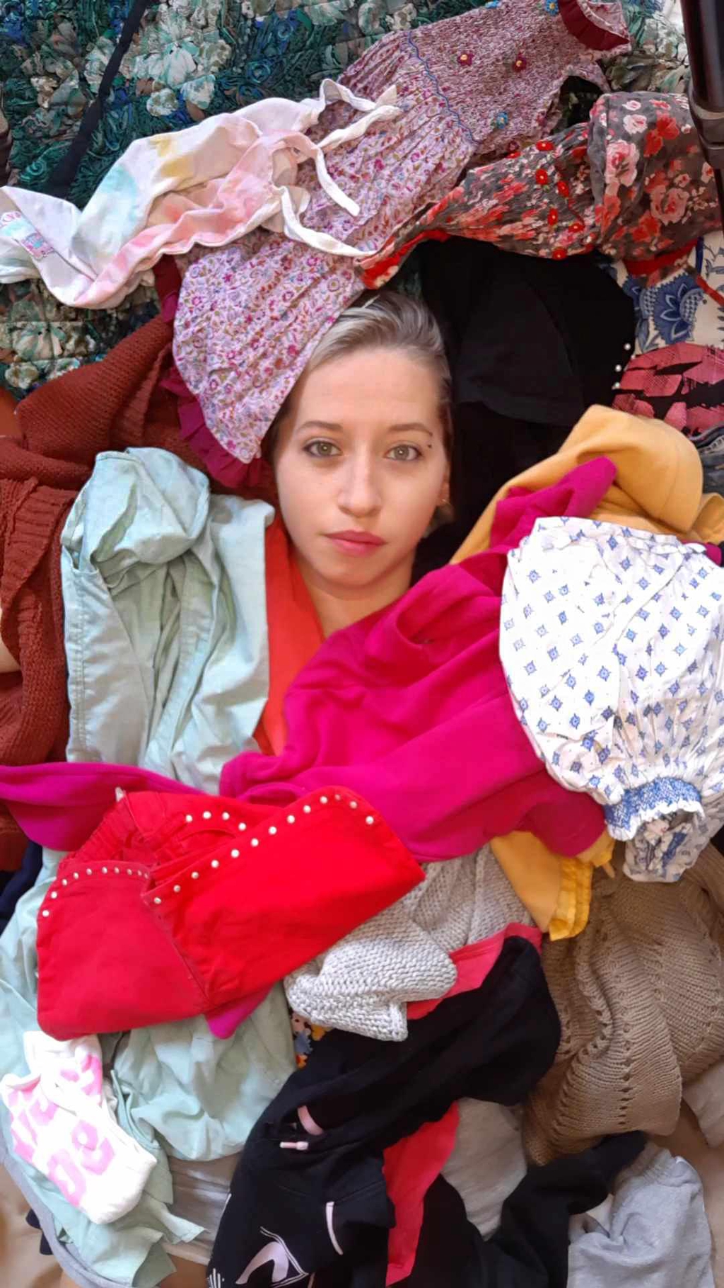La photo représente une femme recouverte d'un tas de vêtement. On ne voit que son visage