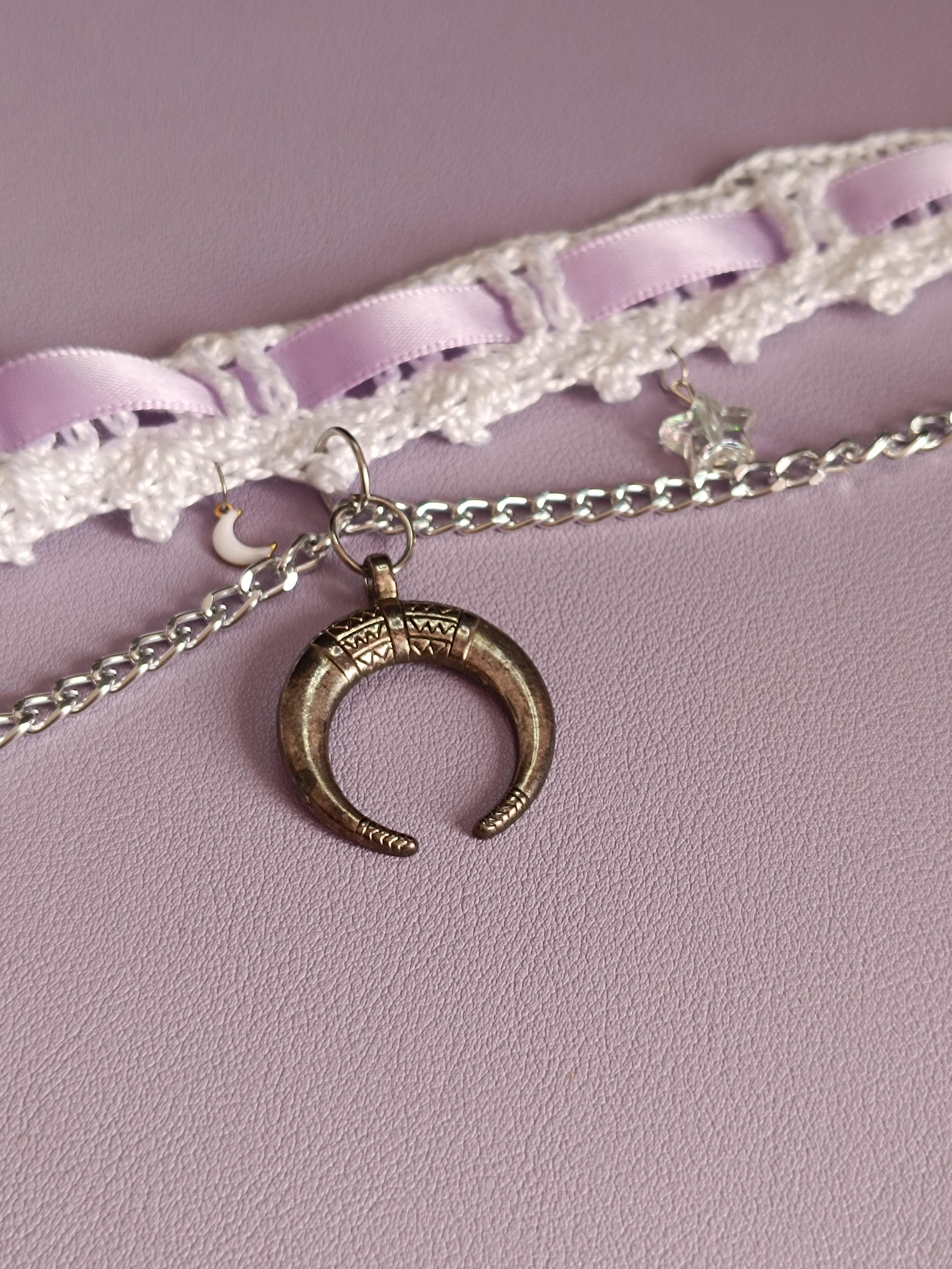 un choker en crochet blanc avec un ruban mauve qui le traverse, et un pendentif croissant de lune argenté.