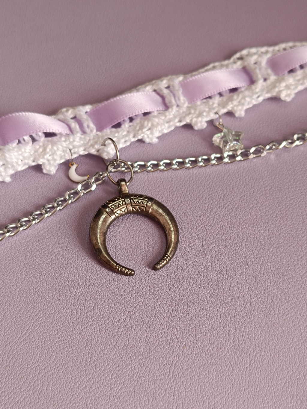 un choker en crochet blanc avec un ruban mauve qui le traverse, et un pendentif croissant de lune argenté.