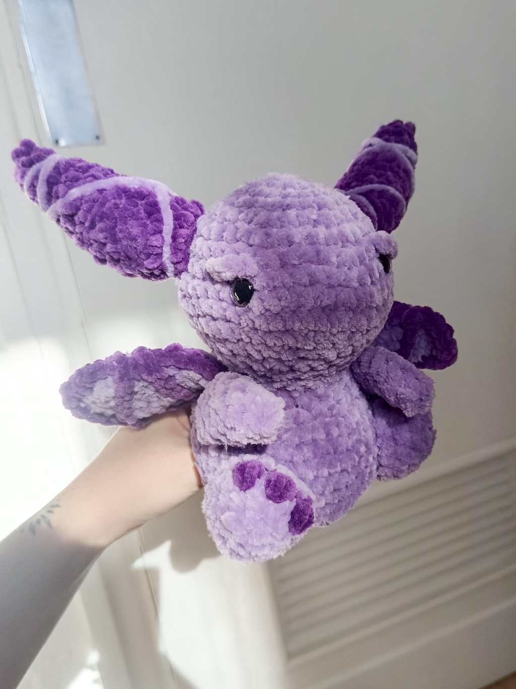 Un dragonnet réalisé en crochet, en laine chenille, il est lilas et ses cornes et ailes sont violettes.