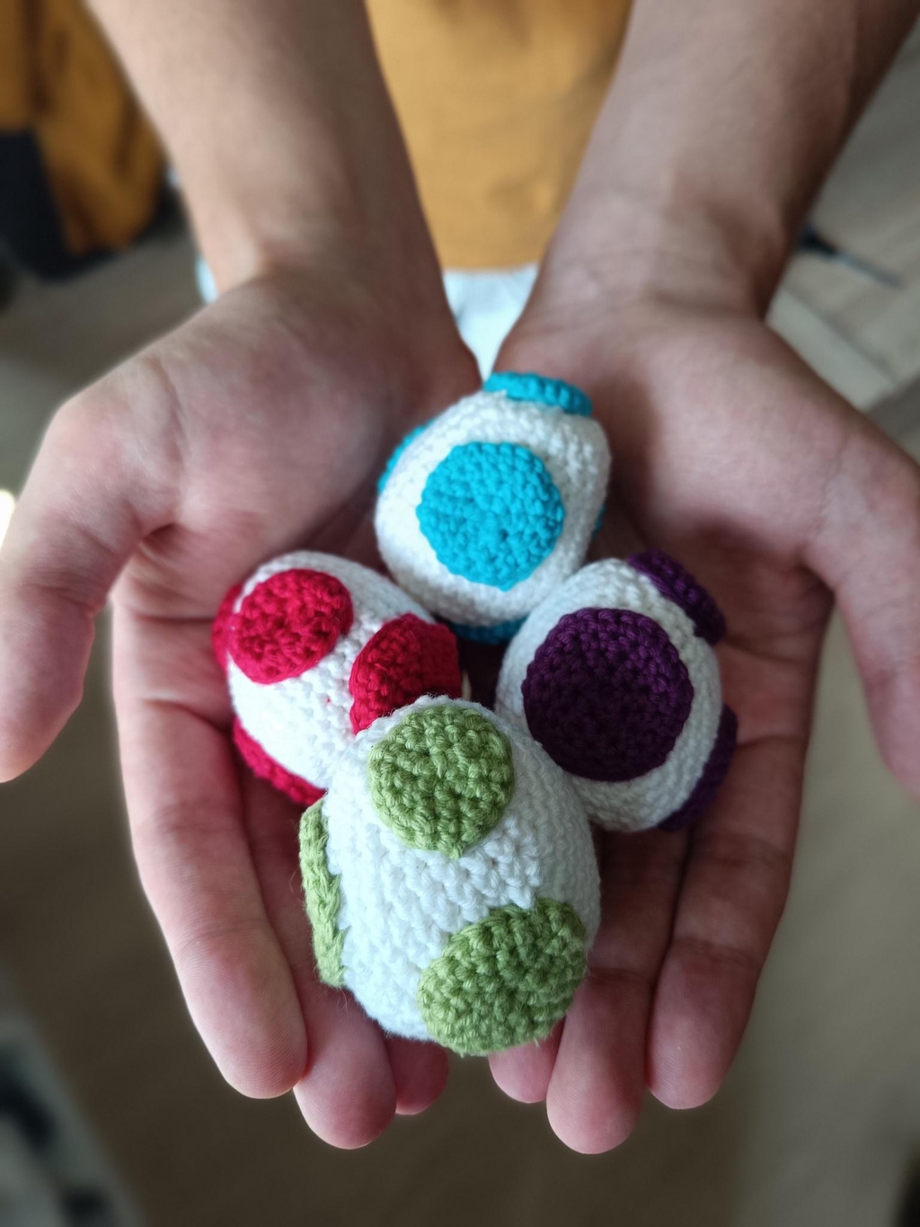 Des oeufs de Yoshi, rouge, bleu, vert et violet, réalisés en crochet, tenues dans deux mains.