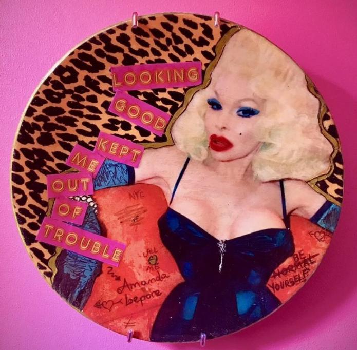 A circular wall plate with a leopard print background, featuring a central figure of a lady with blonde hair in a blue dress with black straps. The artwork includes collage-style text reading 'LOOKING GOOD KEPT ME OUT OF TROUBLE.' Additional handwritten notes, such as 'NYC,' 'ALL,' 'Amanda Lepore,' and 'BE YOURSELF,' are scattered across the piece, adding layers of storytelling and personality. Homage to Amanda Lepore.
