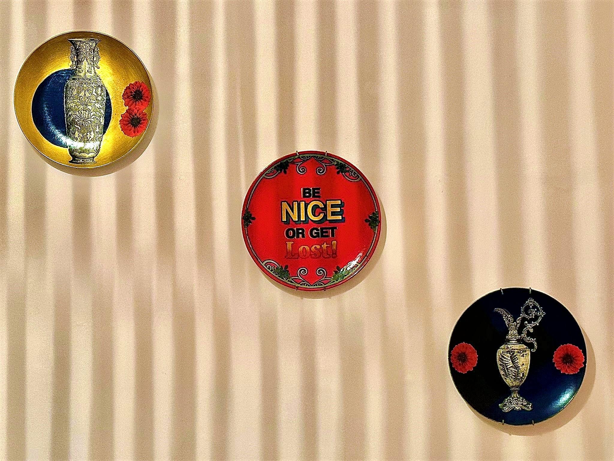 Three decorative plates mounted on a wall arranged in a triangular layout. The top plate features a golden background with an intricate vase design and two red flowers. The bottom left plate has a bold red background with the text 'BE NICE OR GET LOST!' in striking, bold letters and a decorative border. The bottom right plate showcases a dark blue background with an ornate vase design and two red flowers. The background behind the plates has a striped pattern created by shadows or lighting, adding depth to the display. All three decorative wall plates are collage artworks from House of Frisson.