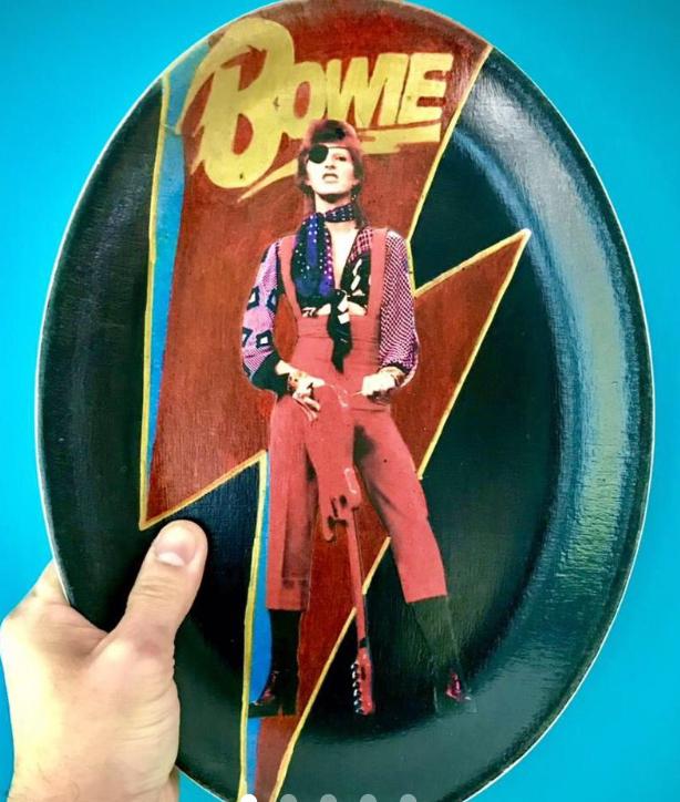 A hand holding an oval-shaped plate featuring a colorful design of David Bowie. The plate displays Bowie in a striking outfit, set against a black background with a bold red and yellow lightning bolt. The word 'Bowie' is prominently displayed in yellow at the top, emphasizing the iconic musician.