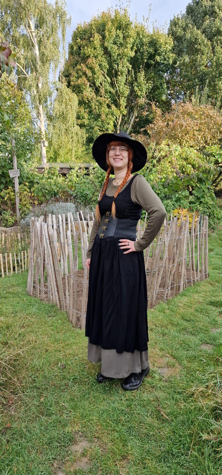 Johanna, Inhaberin von MooonDesigns in einem schwarzen langen Kleid mit grünen Ärmeln und einer schwarzen Korsage. Auf eem Kopf einen schwarzen Hexenhut. Sie steht vor einem Staketenzaun. Dahinter Pflanzen und Büsche.