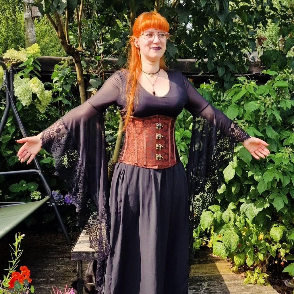 Johanna, Inhaberin von MooonDesigns in einem schwarzen langen Kleid mit einer rotbraunen Korsage. Sie steht in ihrem mittelalterlichen Hexenoutfit umgeben von Pflanzen und Büschen.