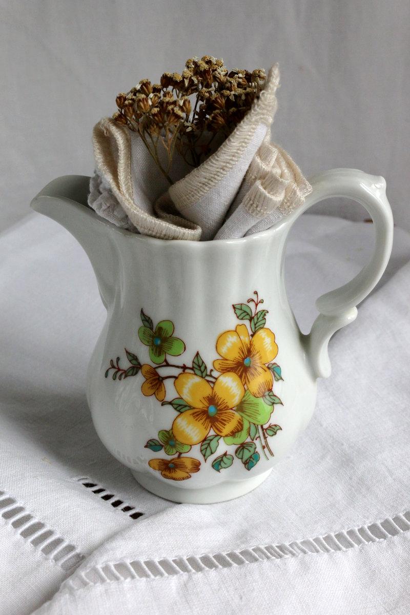 Pichet ancien décoré de motifs floraux jaune et vert contenant une serviette en lin ancien créée à l’atelier, présenté à côté du texte Le concept.