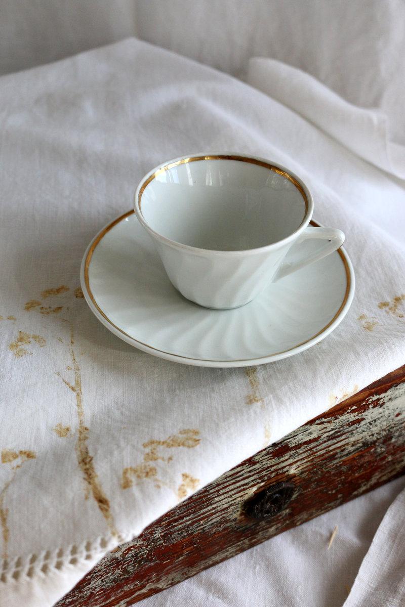 Tasse Félicie De Chauvigny en porcelaine blanche avec anse au filet doré estompé et soucoupe assortie, posées sur un échantillon tatakizome sur plateau en bois brut.