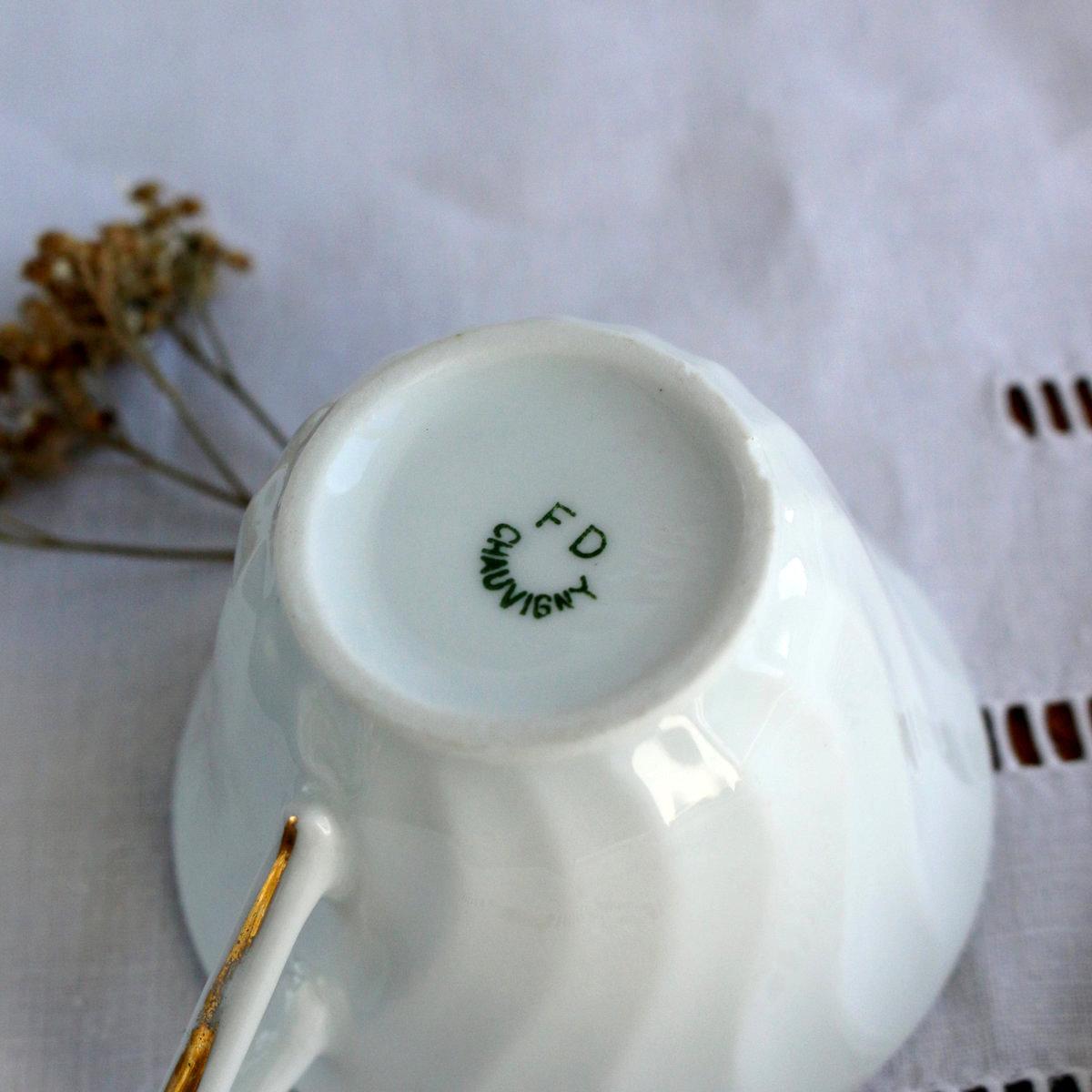 Tasse en porcelaine ancienne retournée montrant le tampon vert F.D. Chauvigny, posée sur une nappe blanche en lin ancien avec jours.