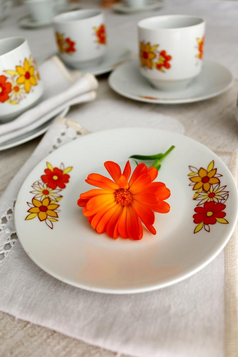 Assiette Winterling Bavaria aux motifs floraux orange et jaune avec une fleur orange posée au centre, sur nappe blanche, avec vaisselle assortie en arrière‑plan.