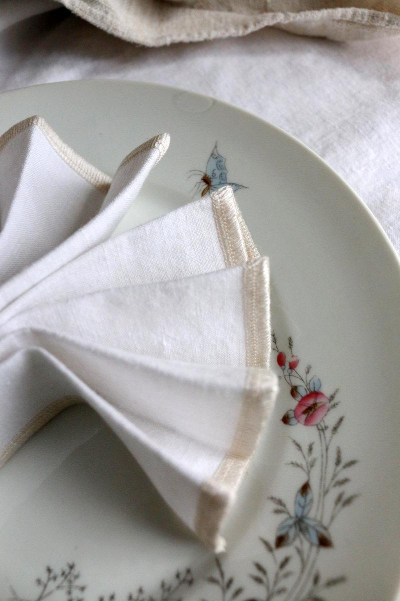 Serviette en lin ancien blanc avec jours et double surjet écru, pliée dans une assiette décorée de fleurs rose, de feuilles légèrement bleues, de tiges brunes et d’un papillon aux ailes bleues.
