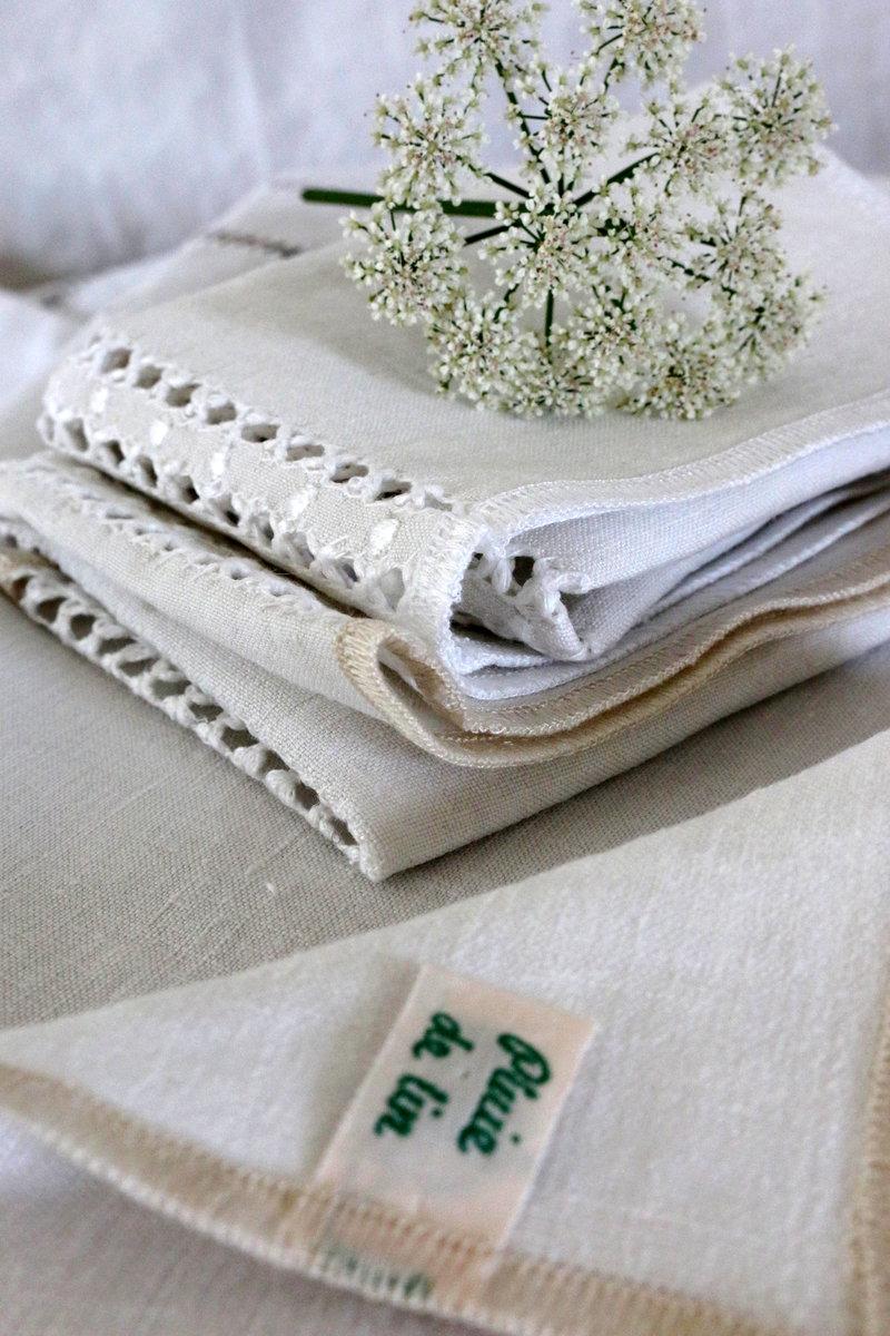 Serviettes en lin ancien blanc avec jours, finition double surjet blanc et écru, posées en pile avec une petite branche de fleurs blanches.