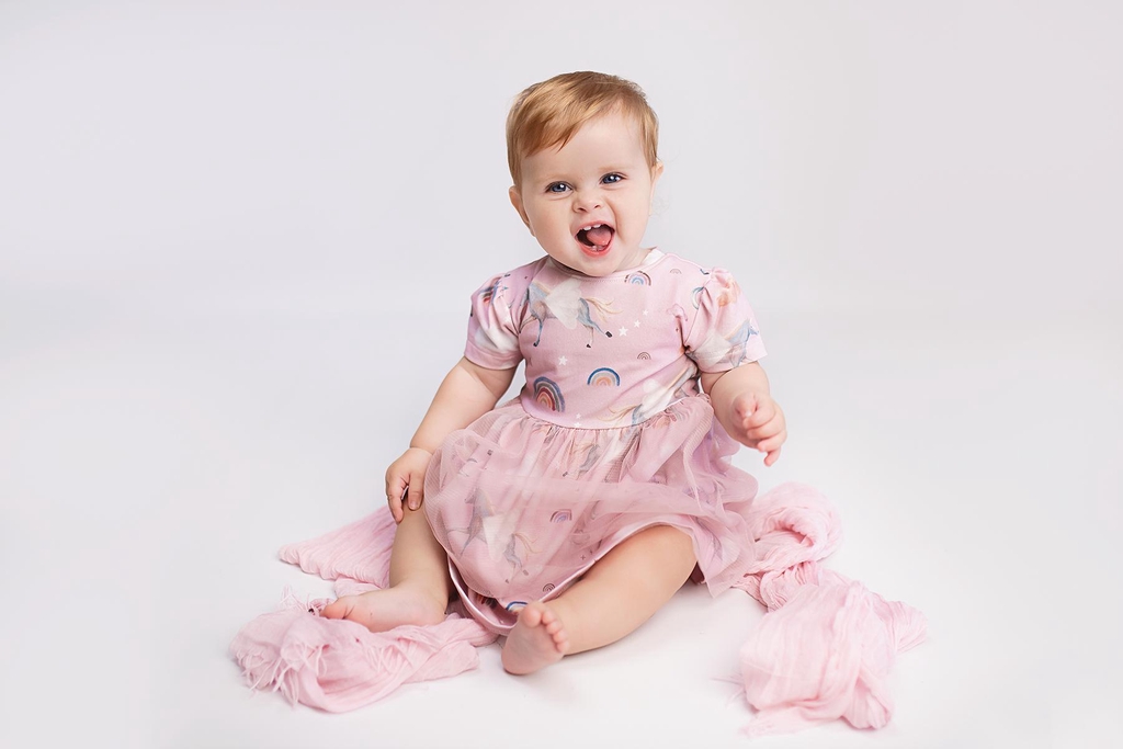 A baby sits in a pink dress with unicorns and tulle. The background is white.