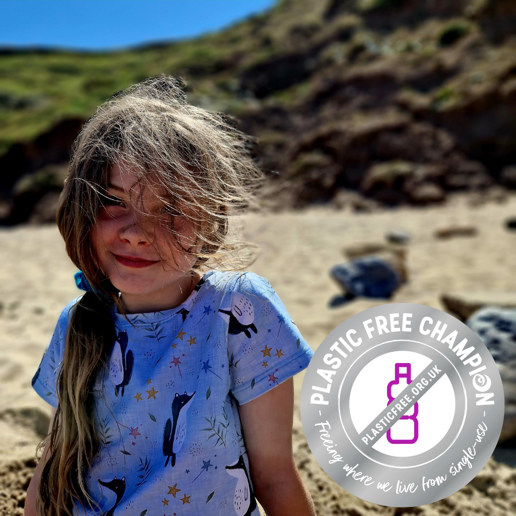 A girl smiles at the camera. She is sat on the beach and wears a Lolabear t shirt in the badgers print. A plastic free champion logo sits to the right of the image.