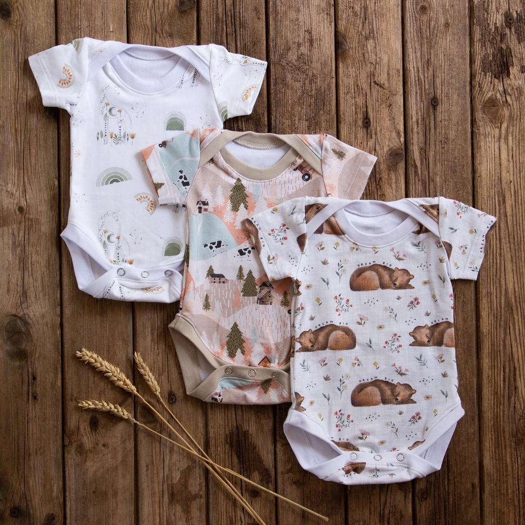 Three baby vests are laid on a wooden background, one with a bear print, one with a farm print and one with a giraffe print.