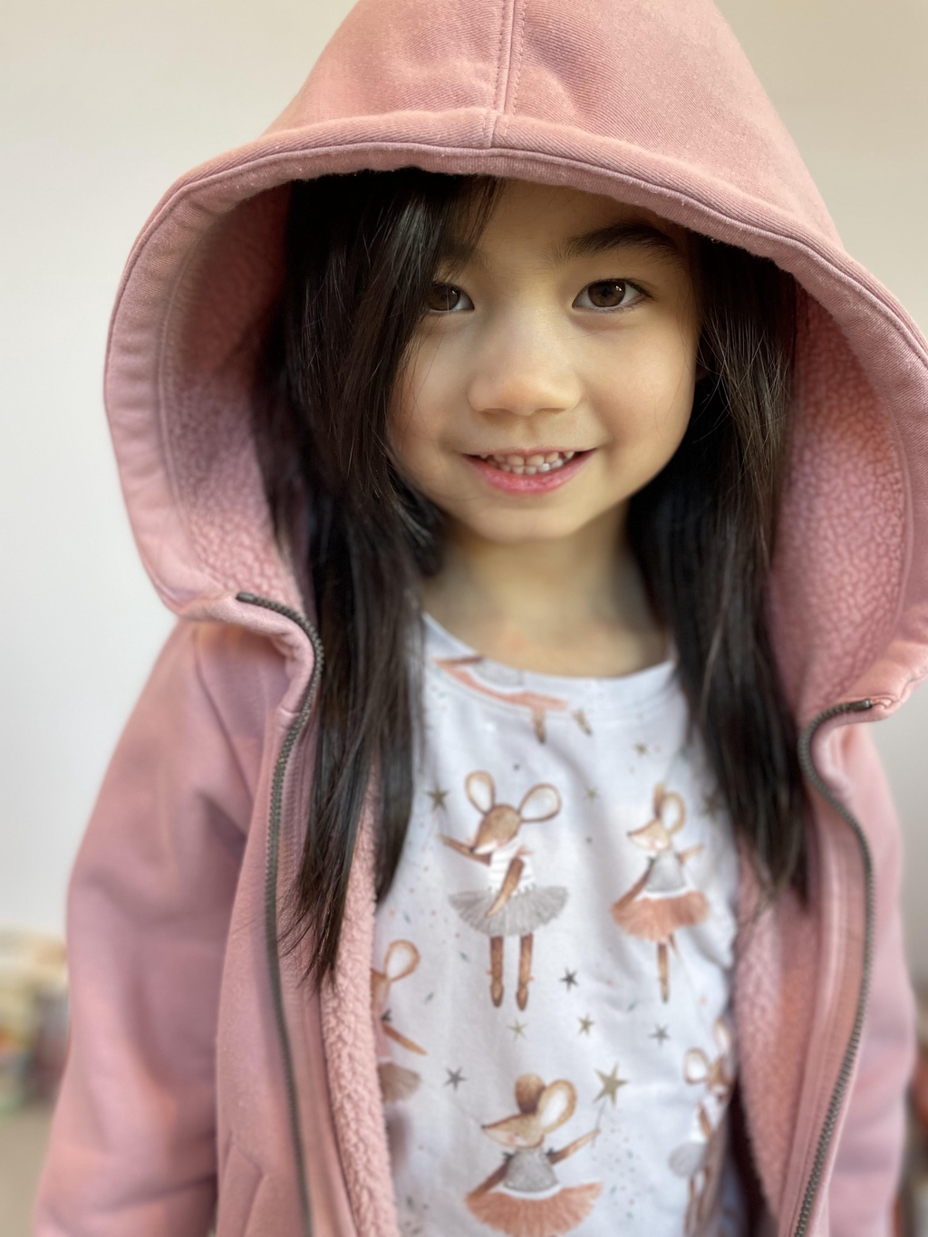 A girl smiles at the camera. She wears a Lolabear ballet mouse t shirts and a pink hoodie with the hood up.