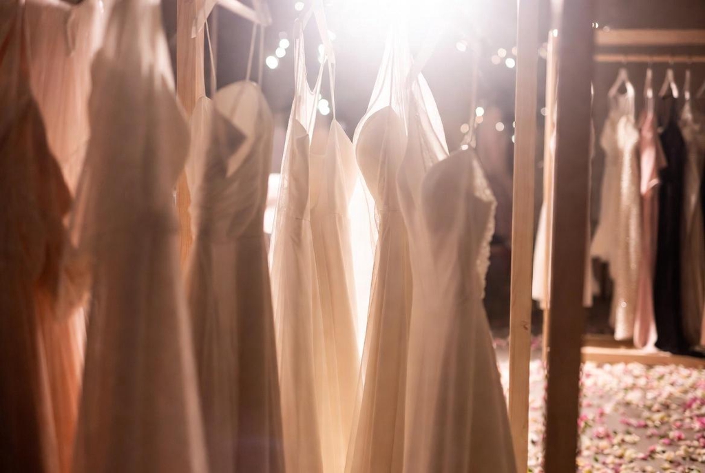 Sélection de robes de mariée simples et joyeuses en tissu fluide ivoire suspendues sur un portant en bois clair avec pétales de fleurs au sol – Collection Maryline Devaux à Fréjus