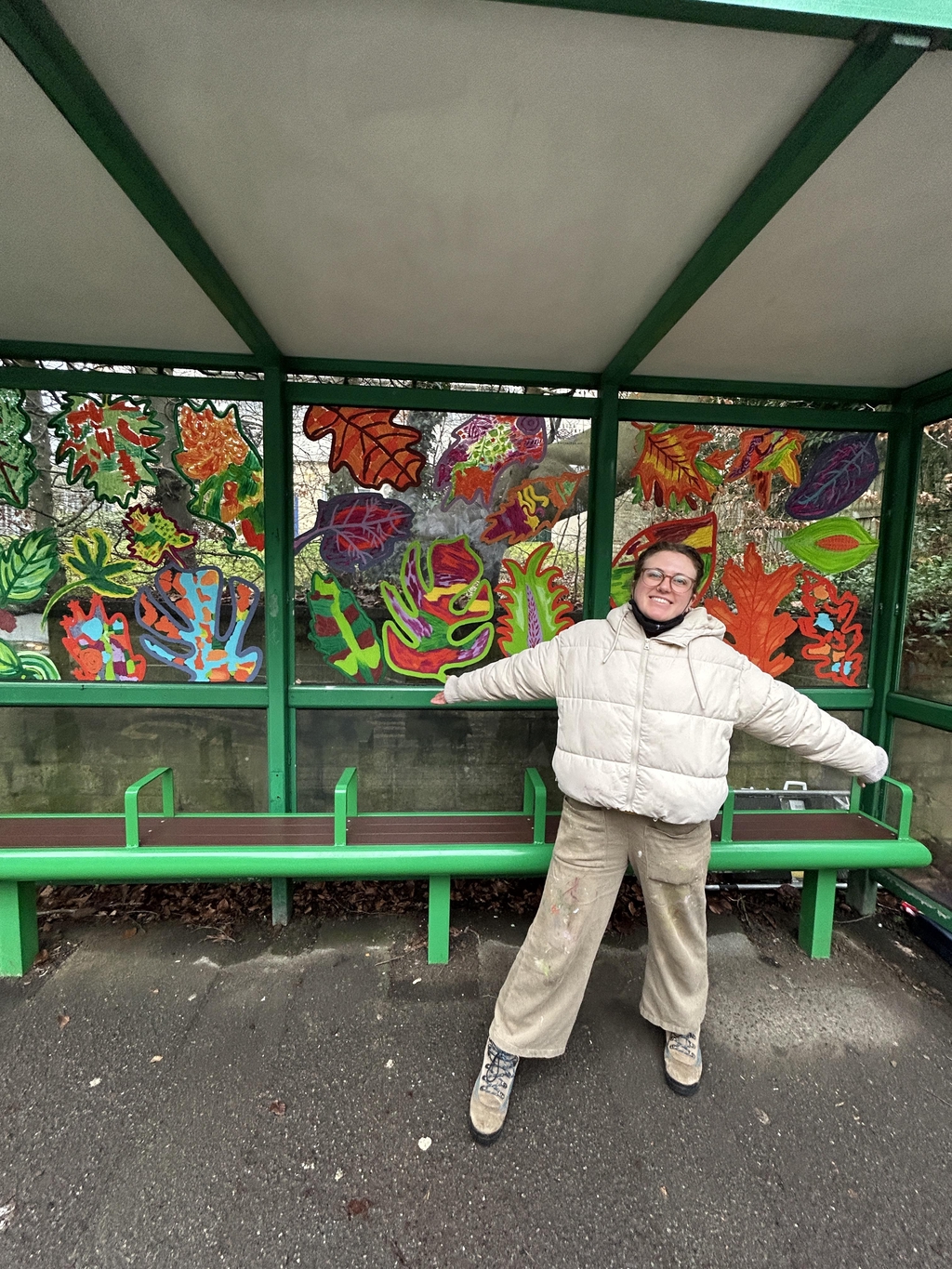 Artist: Eliza skye, stands in front of a mural of colourful bold leaves painted on a green bus stop.
