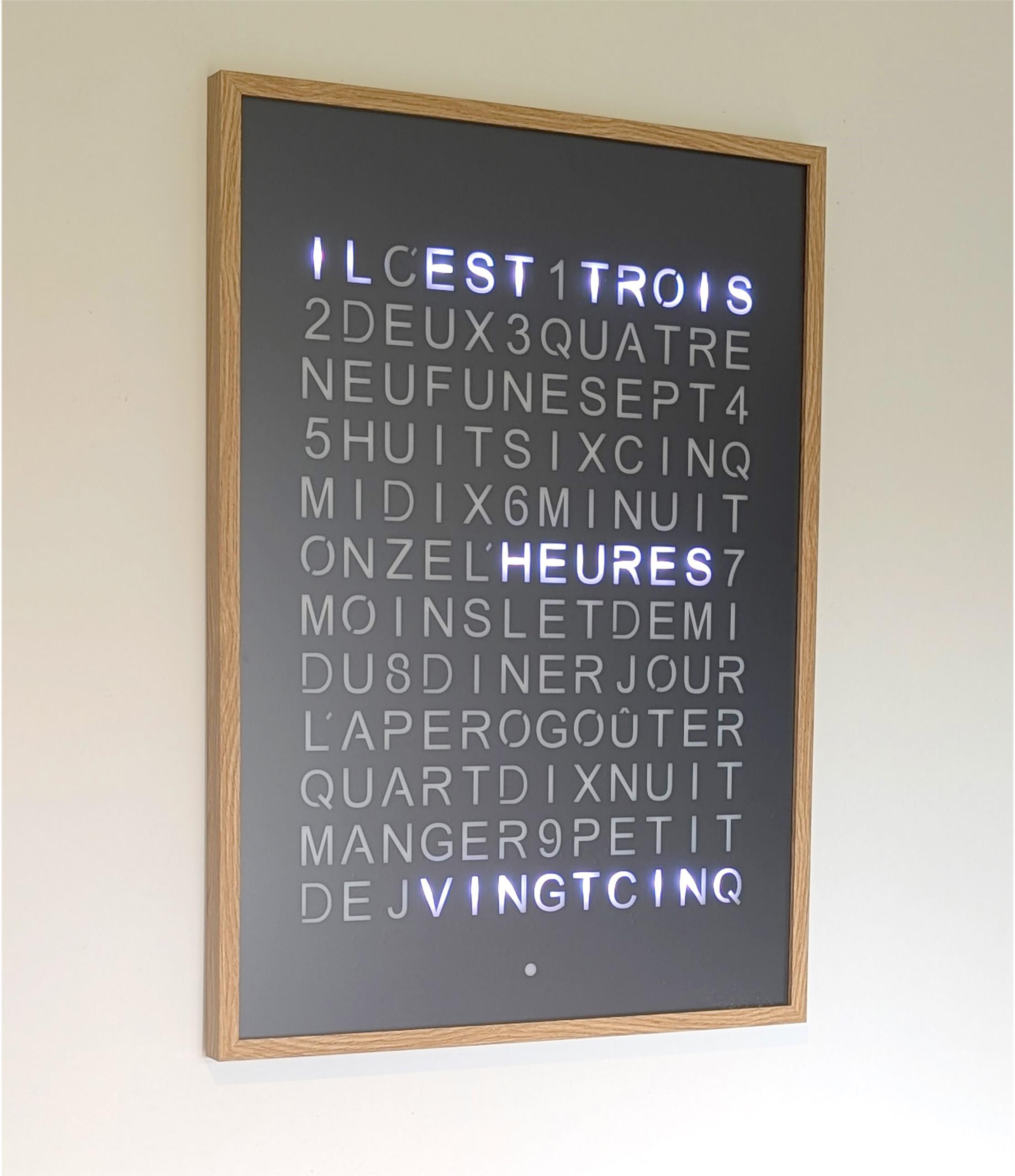 Une horloge à mots murale incorporée sur un cadre de couleur blanc, de couleur bois clair (decor chêne) ou bois foncé (décor noyer) , de dimensions 70 x 50 cm.