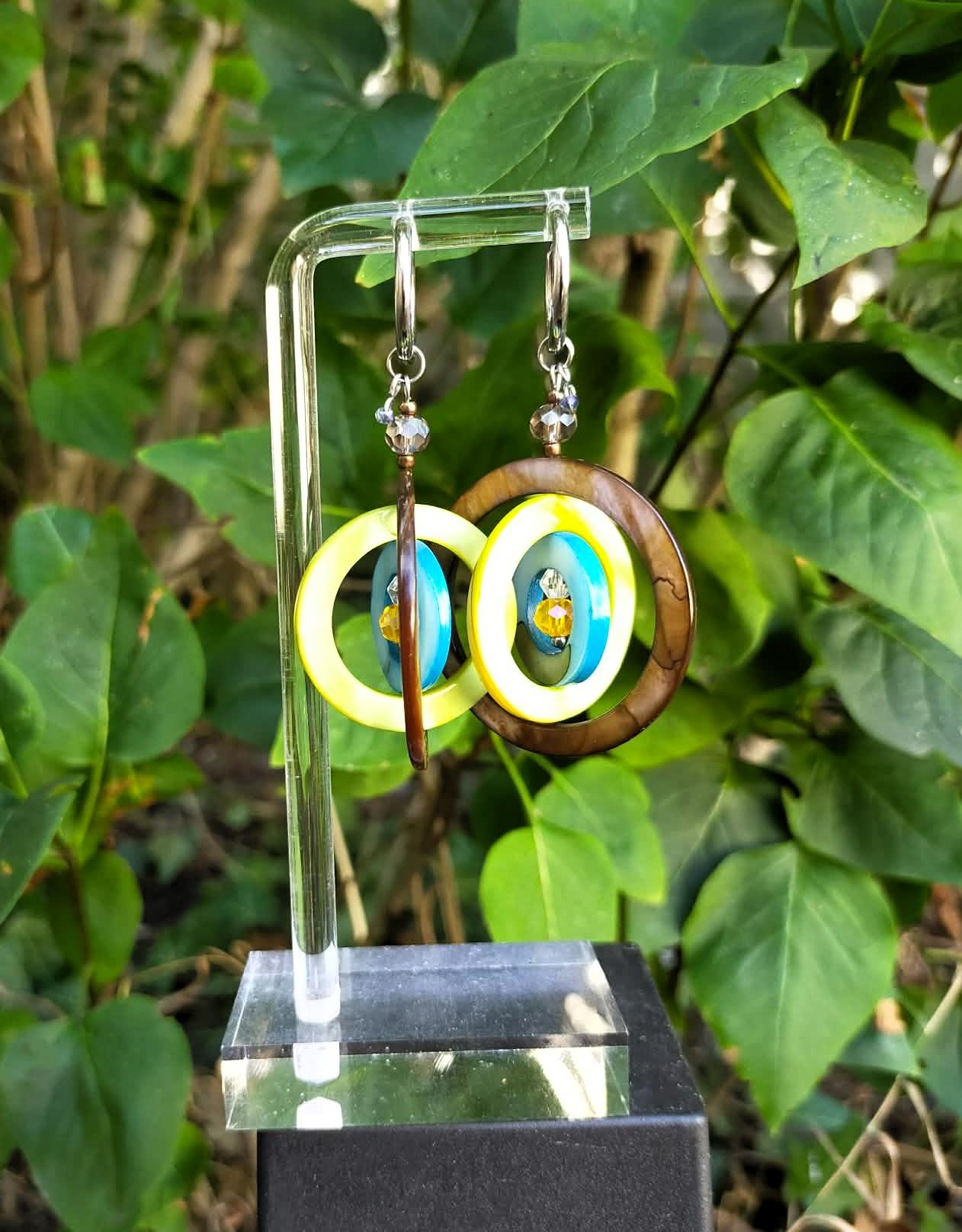Gros plan sur les boucles d'oreilles artisanales Gaïa en nacre et acier inoxydable, présentées sur un support avec fond de feuillage naturel.