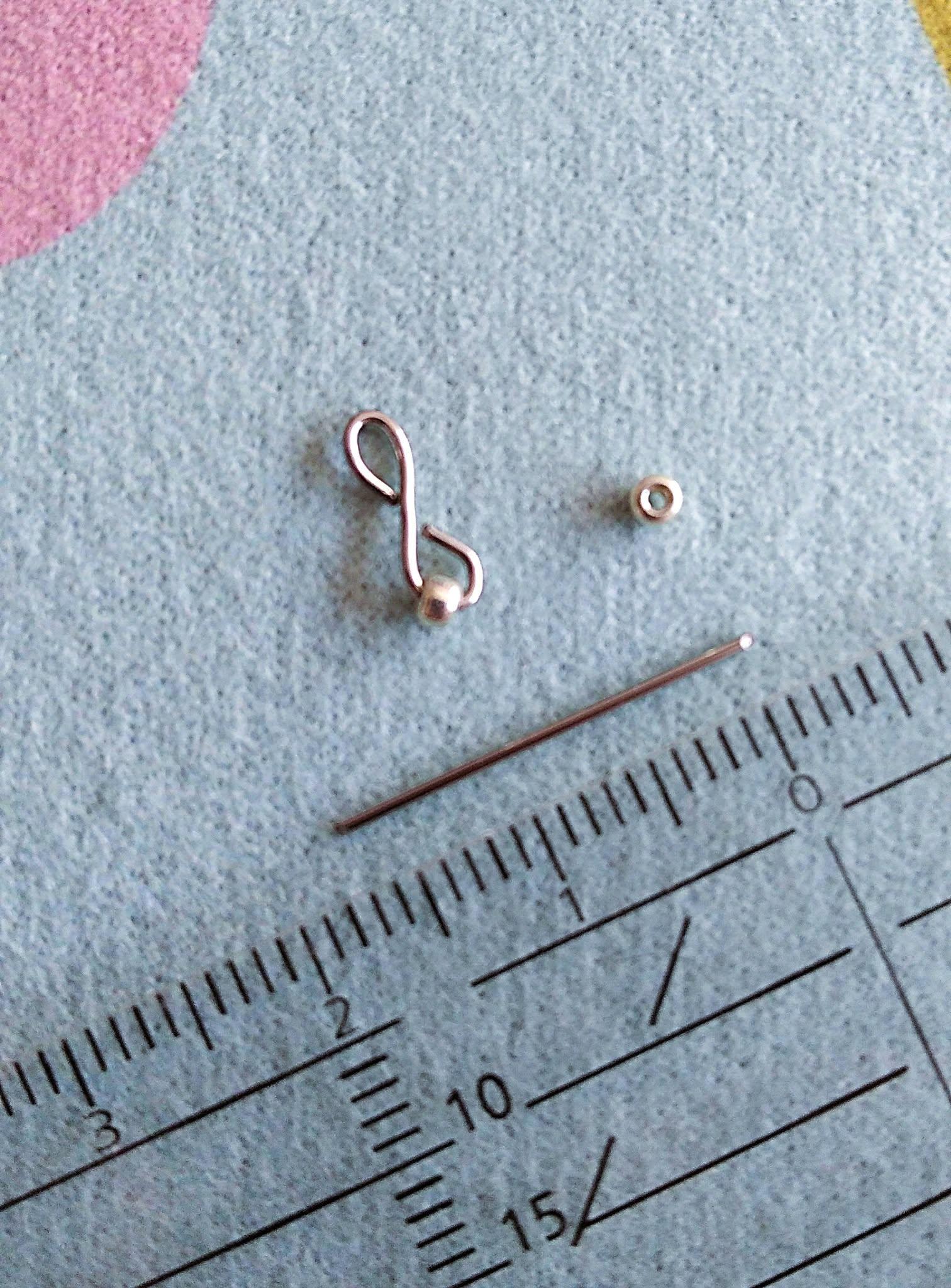 Détail du travail artisanal : symbole de l'infini façonné à la main en acier inoxydable avec mini perle de verre, posé à côté d'une tige et d'une règle sur un tapis de création.