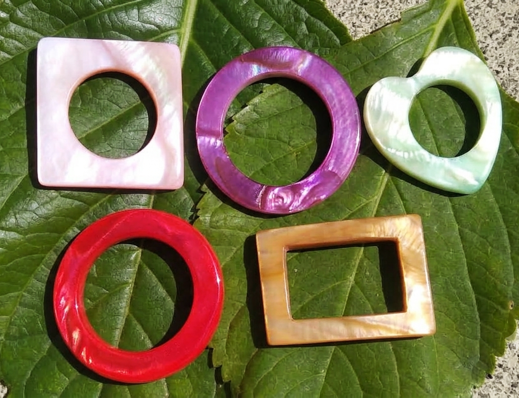 Éléments en nacre naturelle aux reflets irisés colorés posés sur des feuilles vertes, créations de bijoux artisanaux Izia Katell