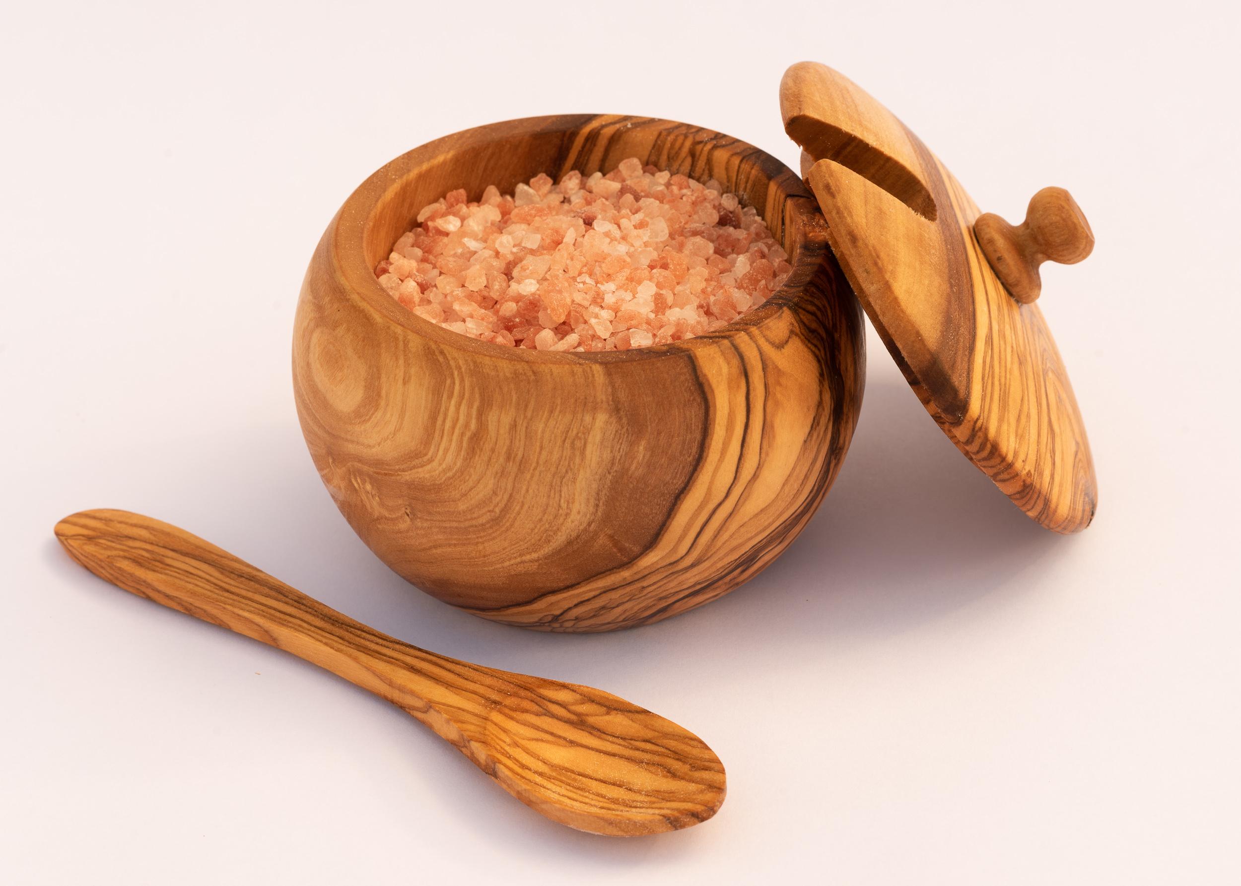 A sugar bowl made from olive wood.  A lid is resting against the bowl and a small olive wood spoon, with rich grains is next to it.  The brightly shot bowl is filled with coarse Himalayan Rock Salt