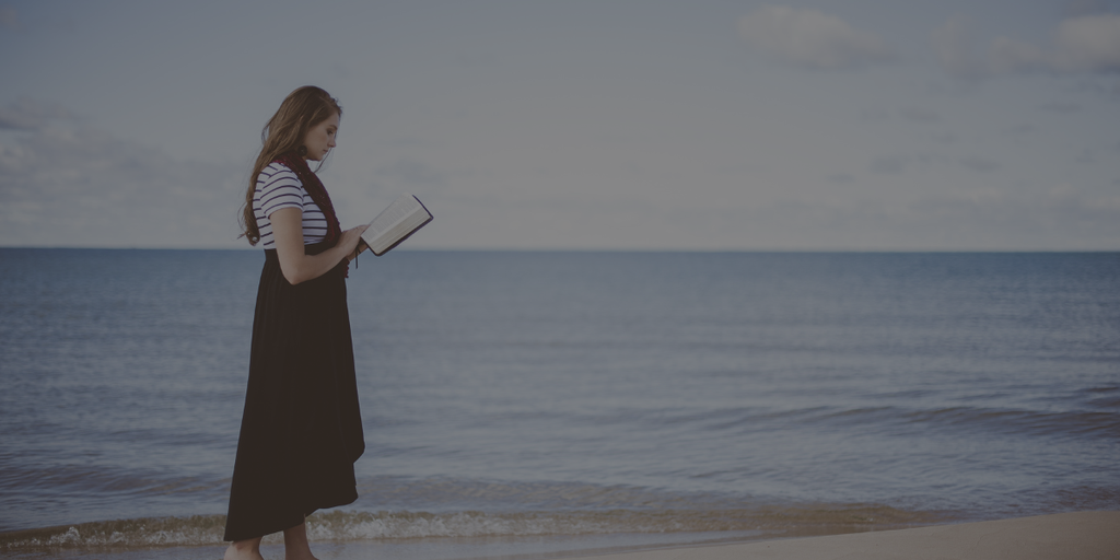 Une jeune femme en robe noire et t-shirt à manches courtes rayé, qui lit un livre debout sur une plage de sable, avec la mer en fond.