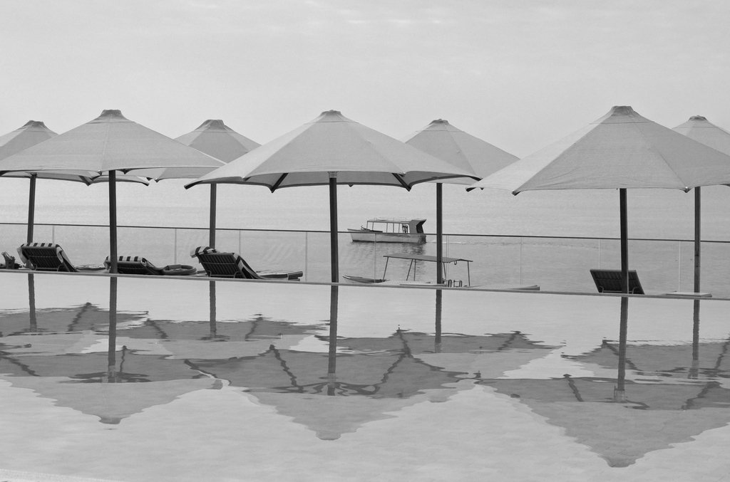 A row of six beach umbrellas photographed in black and white along the edge of an infinity pool overlooking a calm sea. The umbrellas are evenly spaced, their conical canopies mirrored in the still pool water below as diamond-shaped reflections. A few empty lounge chairs sit beneath the umbrellas on the left and right. In the distance, a small boat floats on the flat, hazy water, with a faint horizon line separating sea and sky. Bamburi Beach Resort, Mombasa, Kenya.